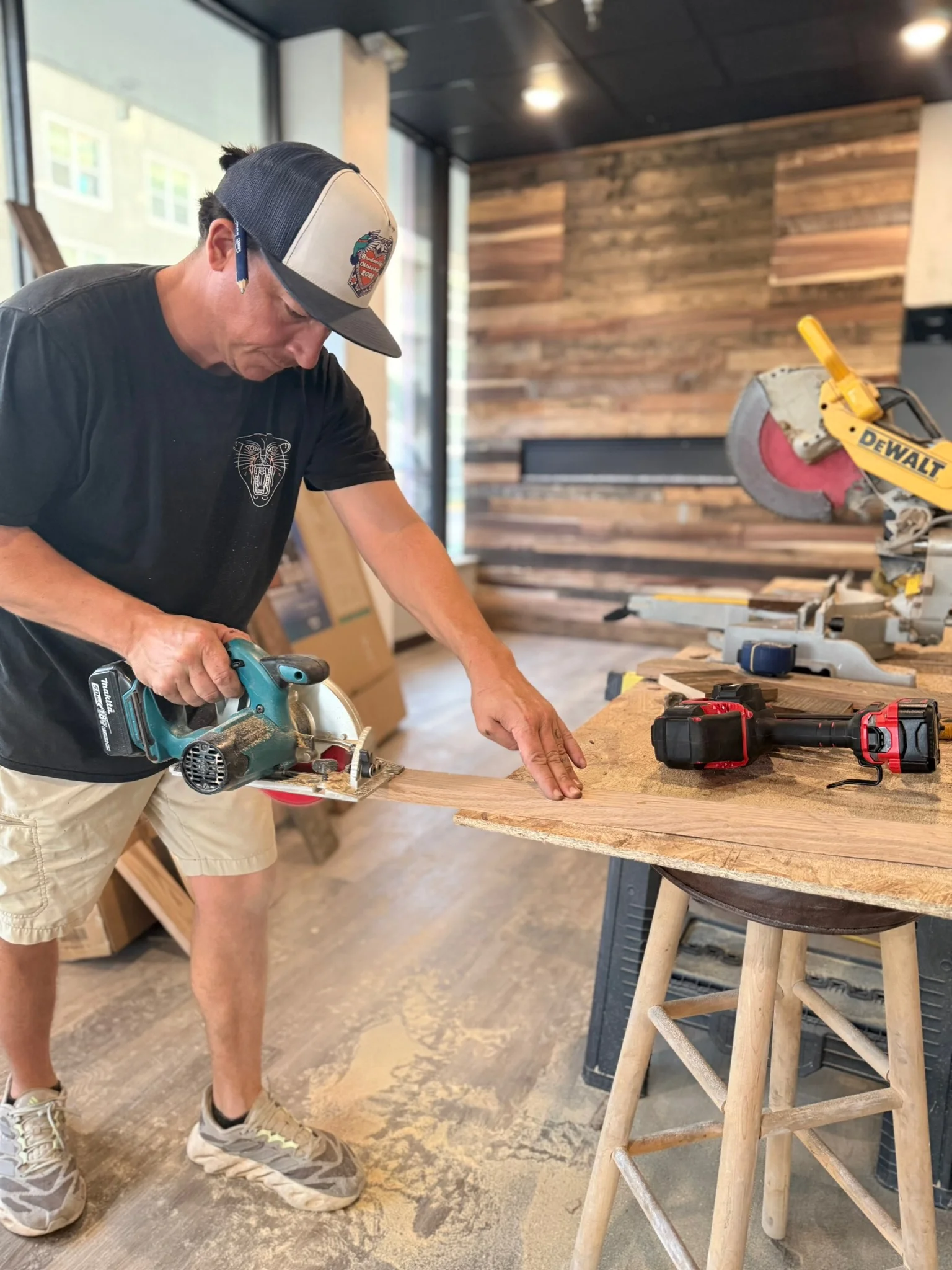 A man is cutting a piece of wood on a workbench with a circular saw inside a woodworking shop. A cordless drill is placed on the workbench next to the wood. The shop has a wooden accent wall and large windows bringing in natural light.