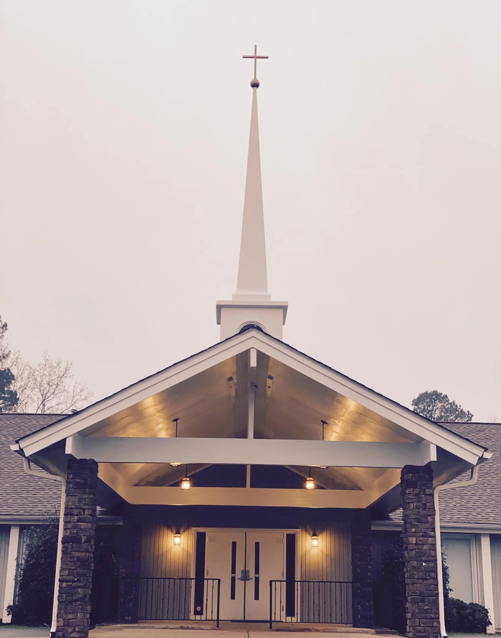Front view of a church with a tall steeple topped with a cross, stone columns on either side of the entrance, and lights illuminating the porch area.