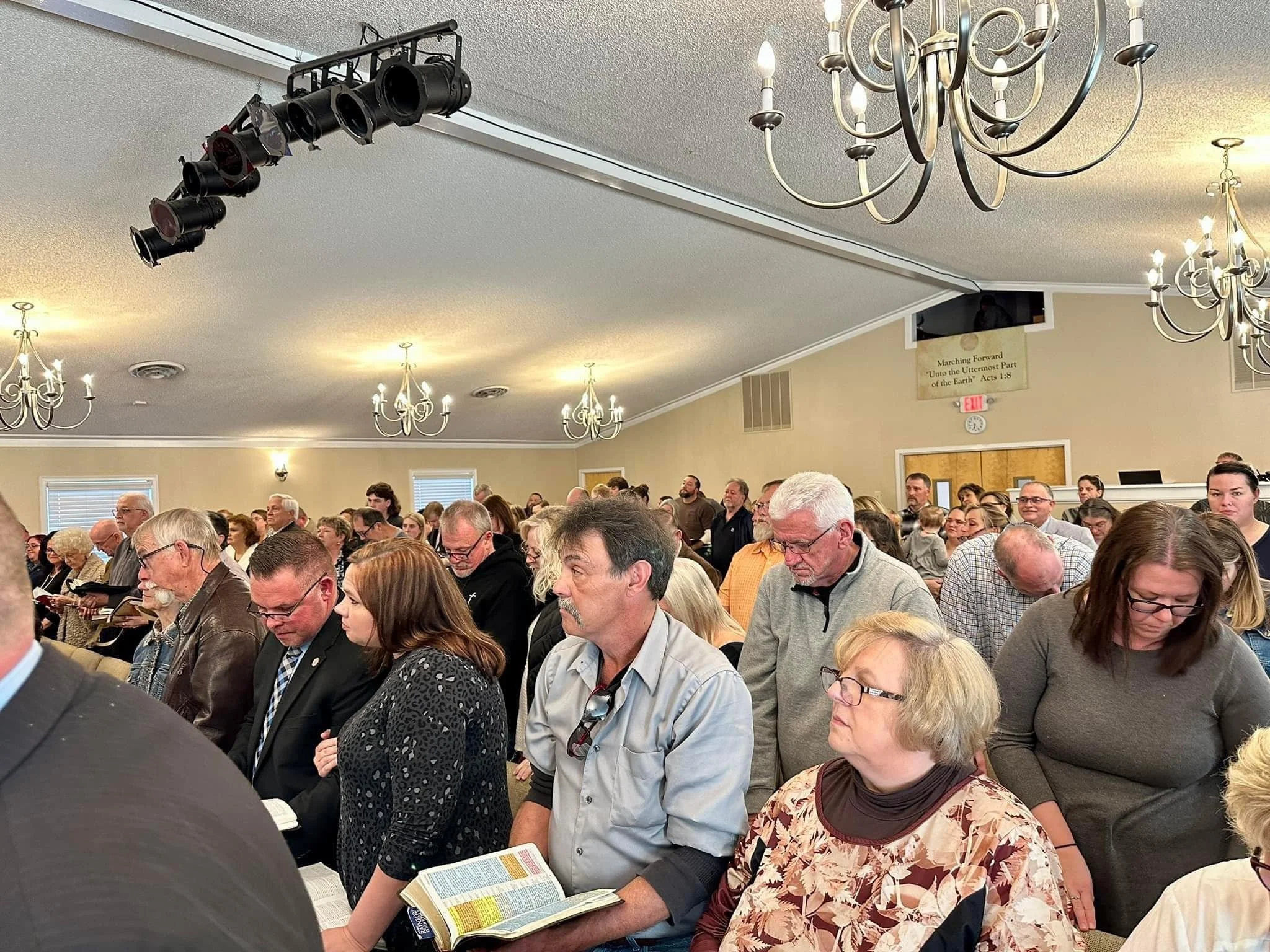 People standing in church pews during a prayer service, many with eyes closed and heads bowed, in a large church with chandeliers.