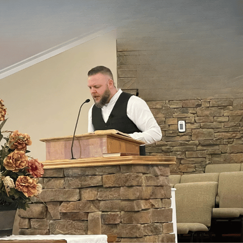 A man in a white shirt and black vest standing at a wooden pulpit with an open book, speaking into a microphone, in a room with a brick and beige wall, and a beige couch in the background.