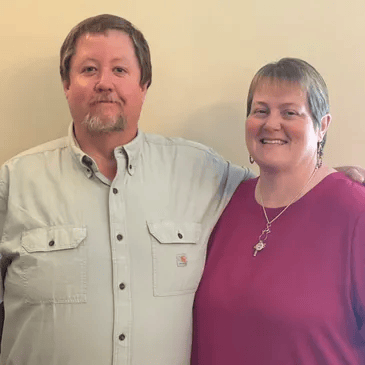 A man and woman standing side by side, smiling, against a plain beige wall.