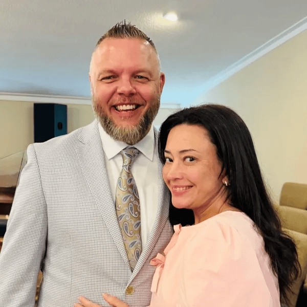 A smiling man with a beard in a light-colored suit and a woman with dark hair in a pink blouse, standing together indoors.