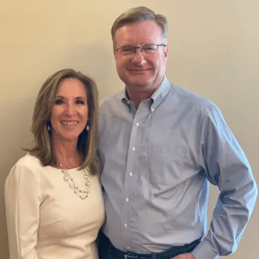 A woman and a man standing side by side, smiling, in front of a plain beige wall.