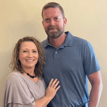 A smiling woman and man posing together against a yellow wall, with the woman wearing a light-colored top and a necklace, and the man wearing a blue polo shirt.