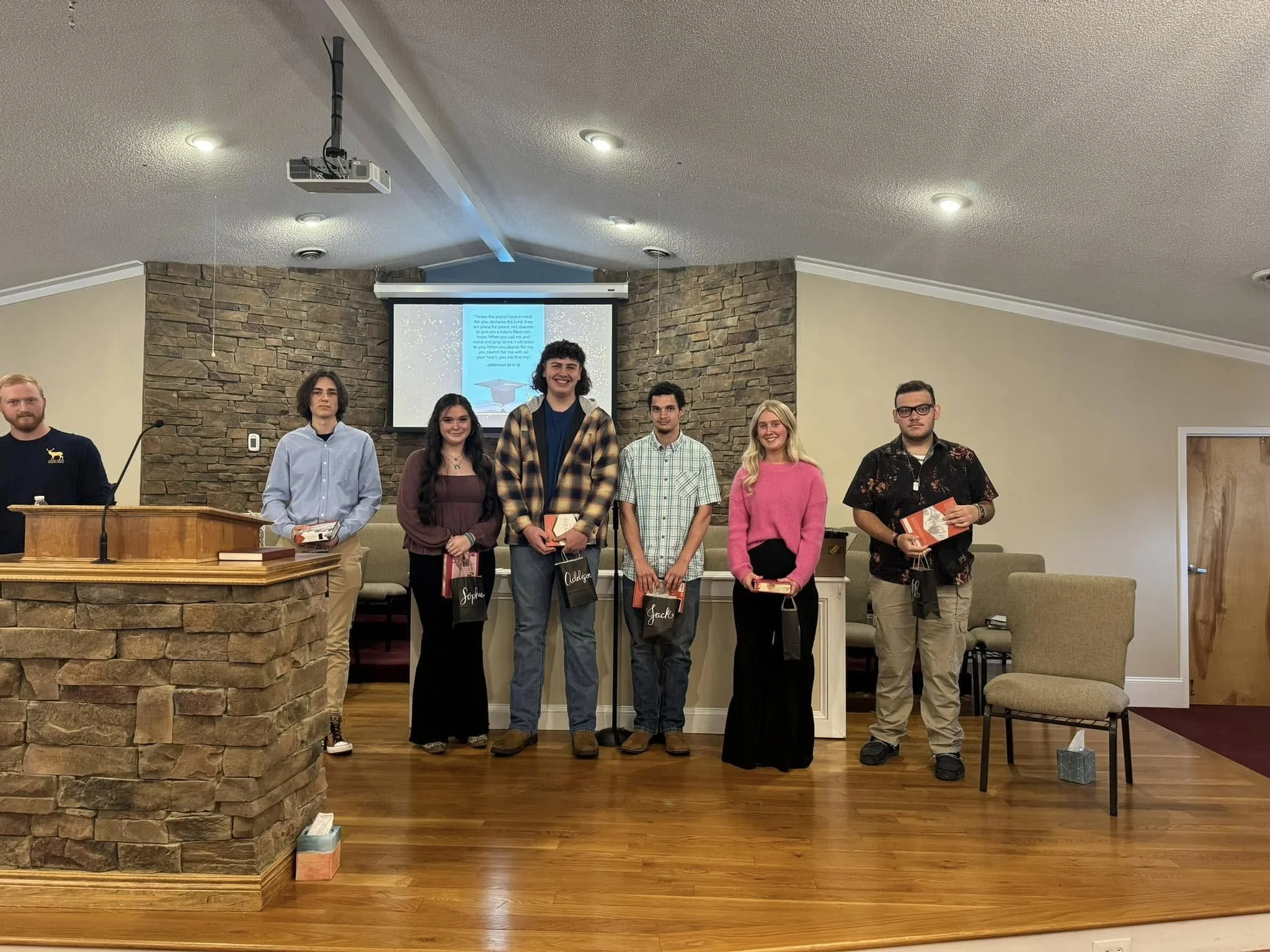 Group of seven people standing on a stage in a church or conference room, some holding books or awards, with a wooden podium, a brick wall, and a screen displaying text and a picture of a graduation cap in the background.