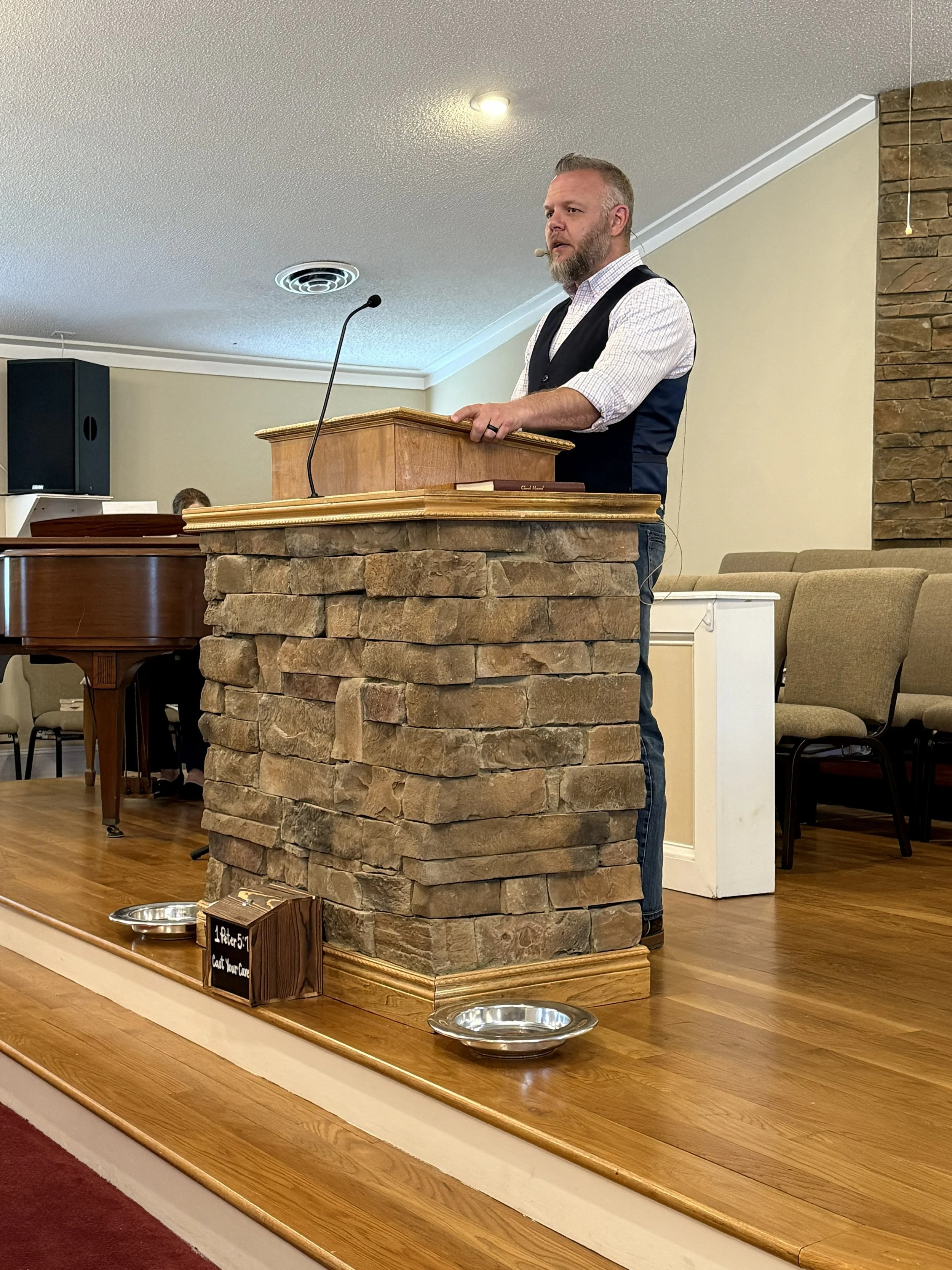 Man in a black vest and speaking at a wooden podium in a room with beige walls and stone accents.