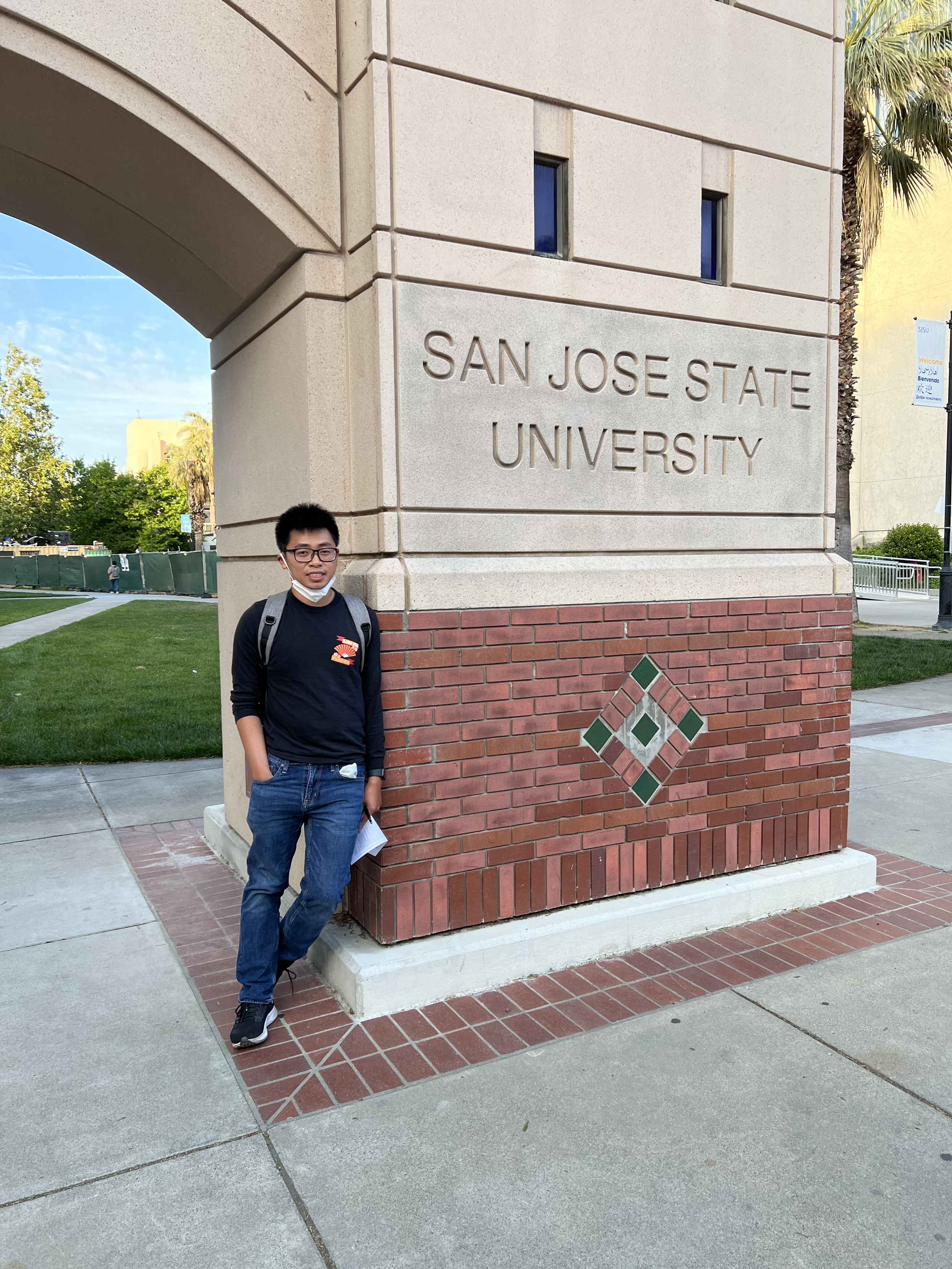 A young man with glasses, wearing a face mask around his neck, a black long-sleeve shirt, and jeans, standing next to a large stone sign that reads "San Jose State University." The sign is on a brick and concrete base, with a decorative diamond pattern at the bottom. In the background, there are trees, a sidewalk, and a building.