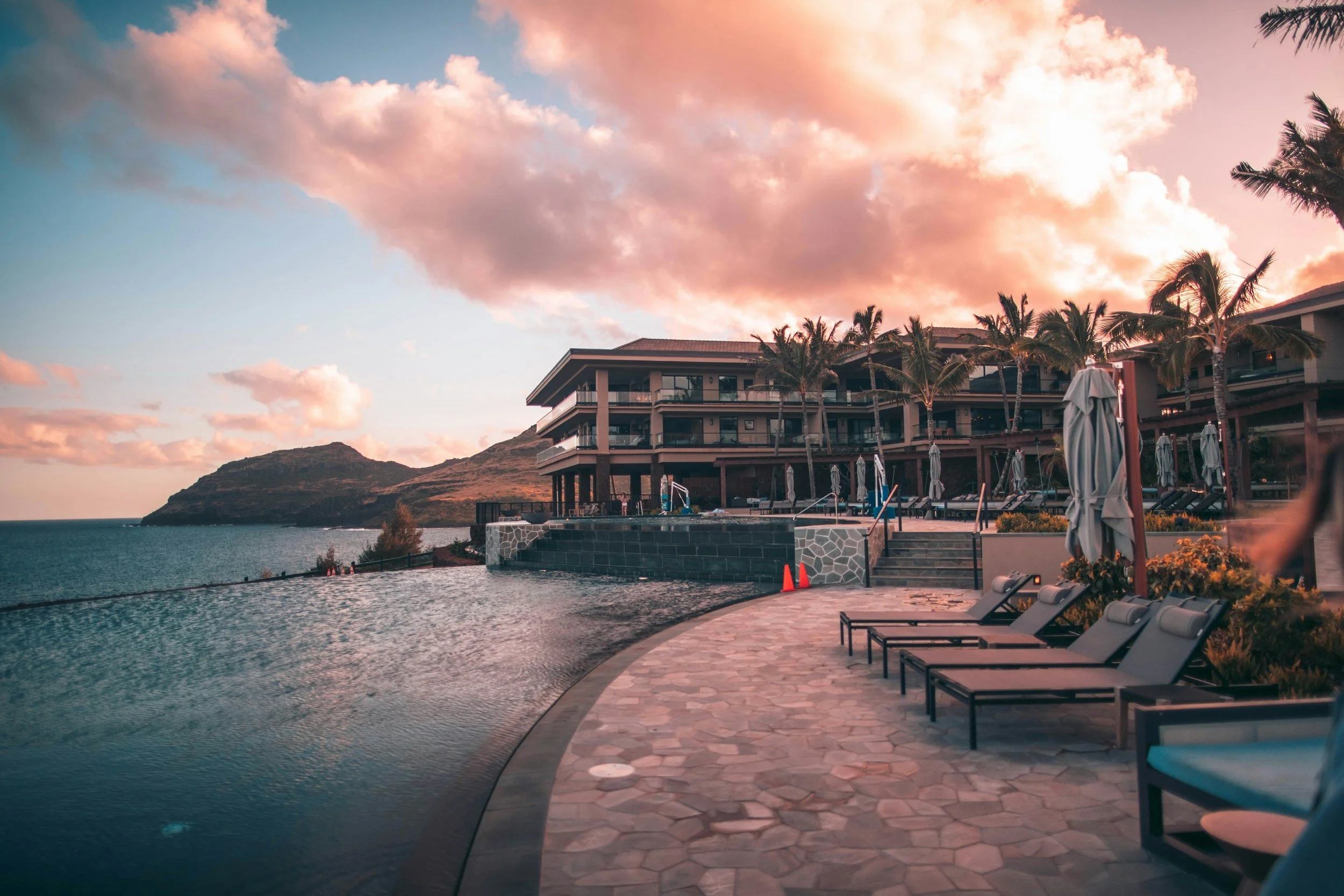 Luxury hotel with a swimming pool, surrounded by palm trees, overlooking a coastal hill at sunset.