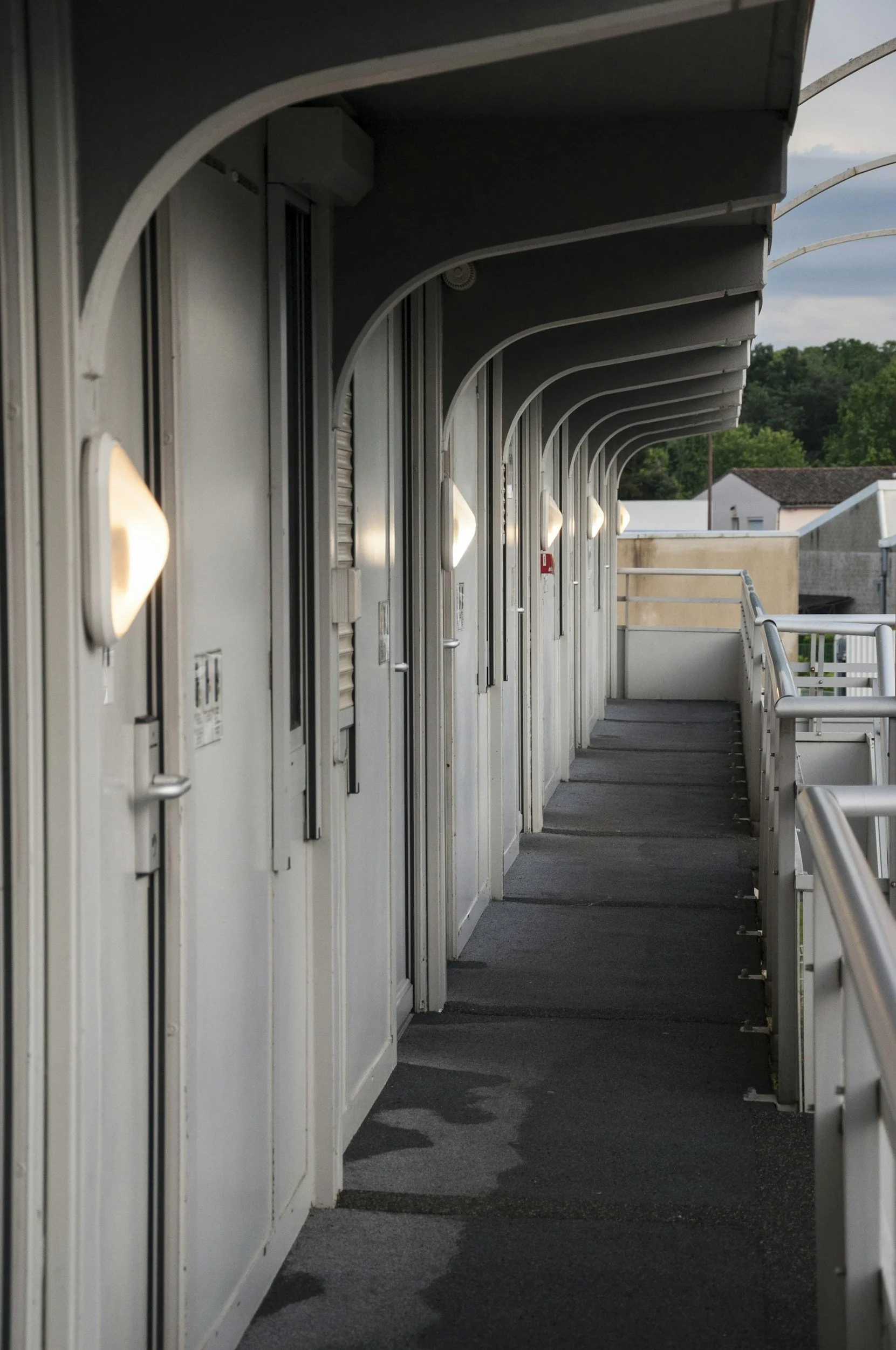 A balcony or walkway with doors on the left side, round light fixtures illuminating the corridor, and a view of trees and buildings in the distance.