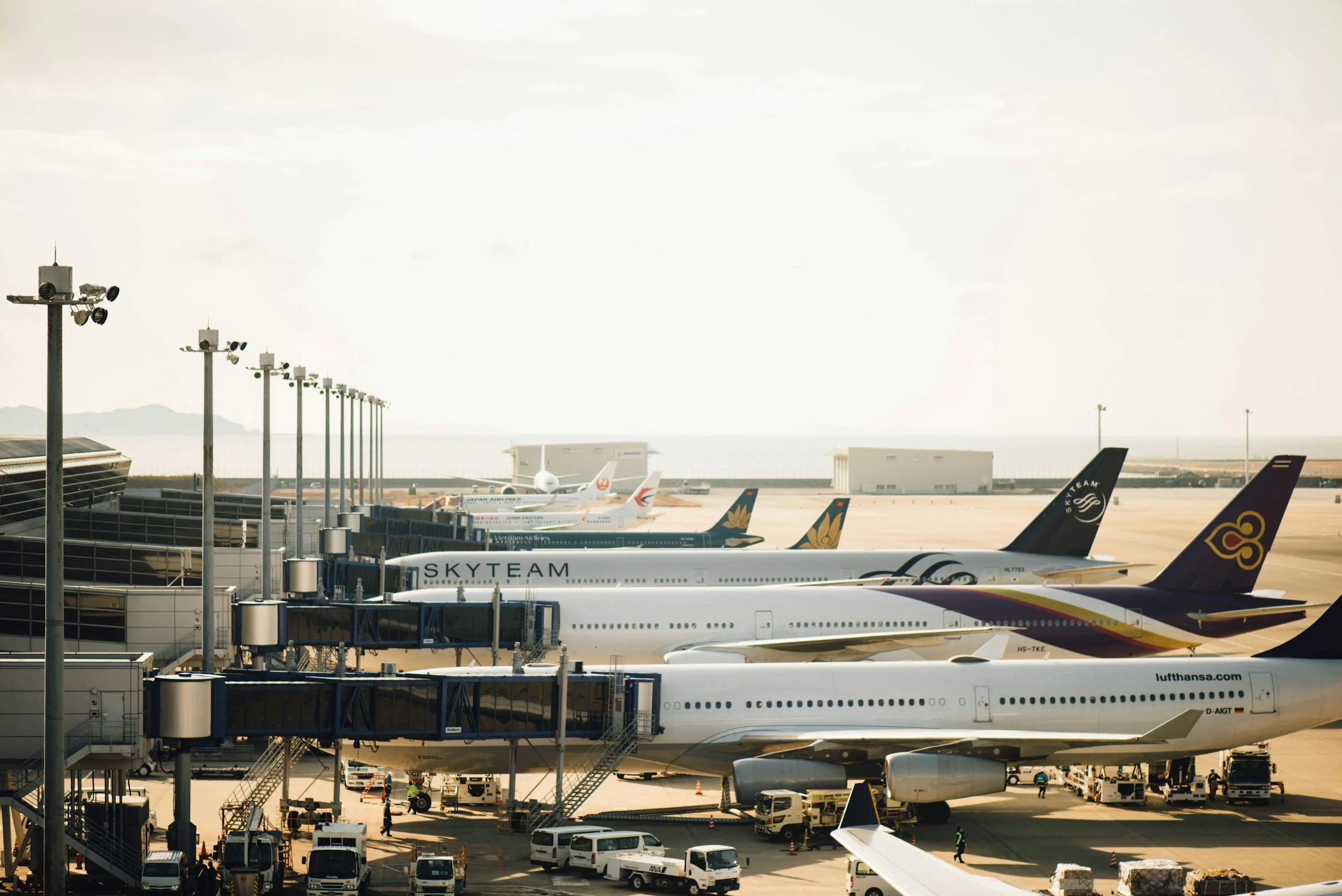 View of an airport terminal with multiple airplanes parked at the gates, including jets with SkyTeam and Lufthansa logos, and ground service vehicles nearby.