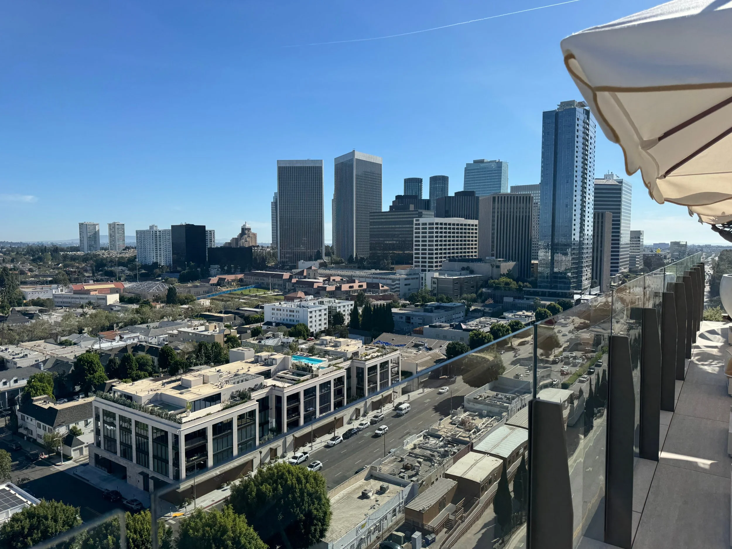 View of a city skyline with tall office buildings and residential structures, under a clear blue sky. An outdoor balcony with a glass railing extends from the foreground, with part of a white patio umbrella visible on the right side.