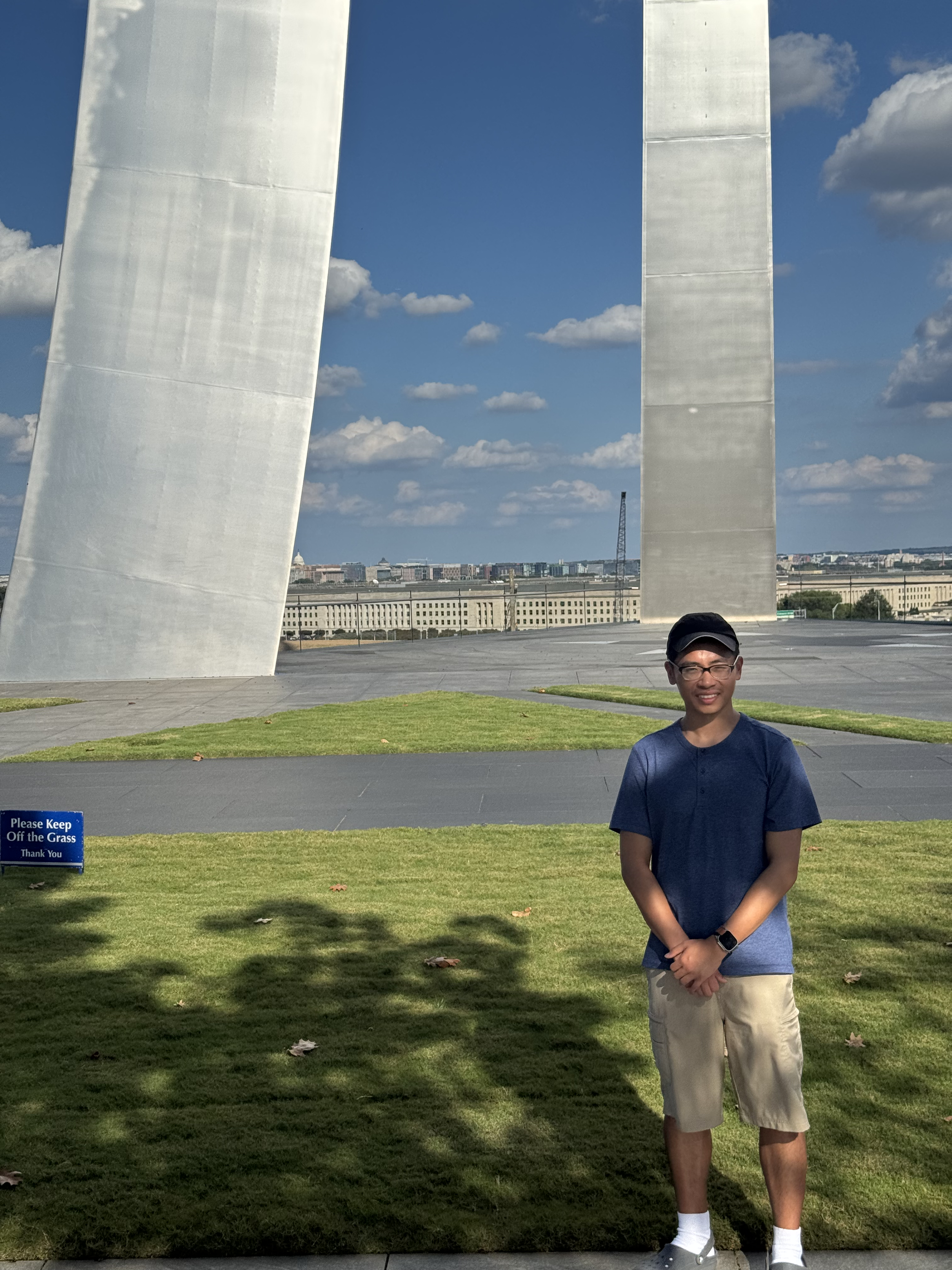 A young man standing on a grassy area in front of the World War II Memorial in Washington, D.C., with the memorial's large steel structures and a city skyline in the background.