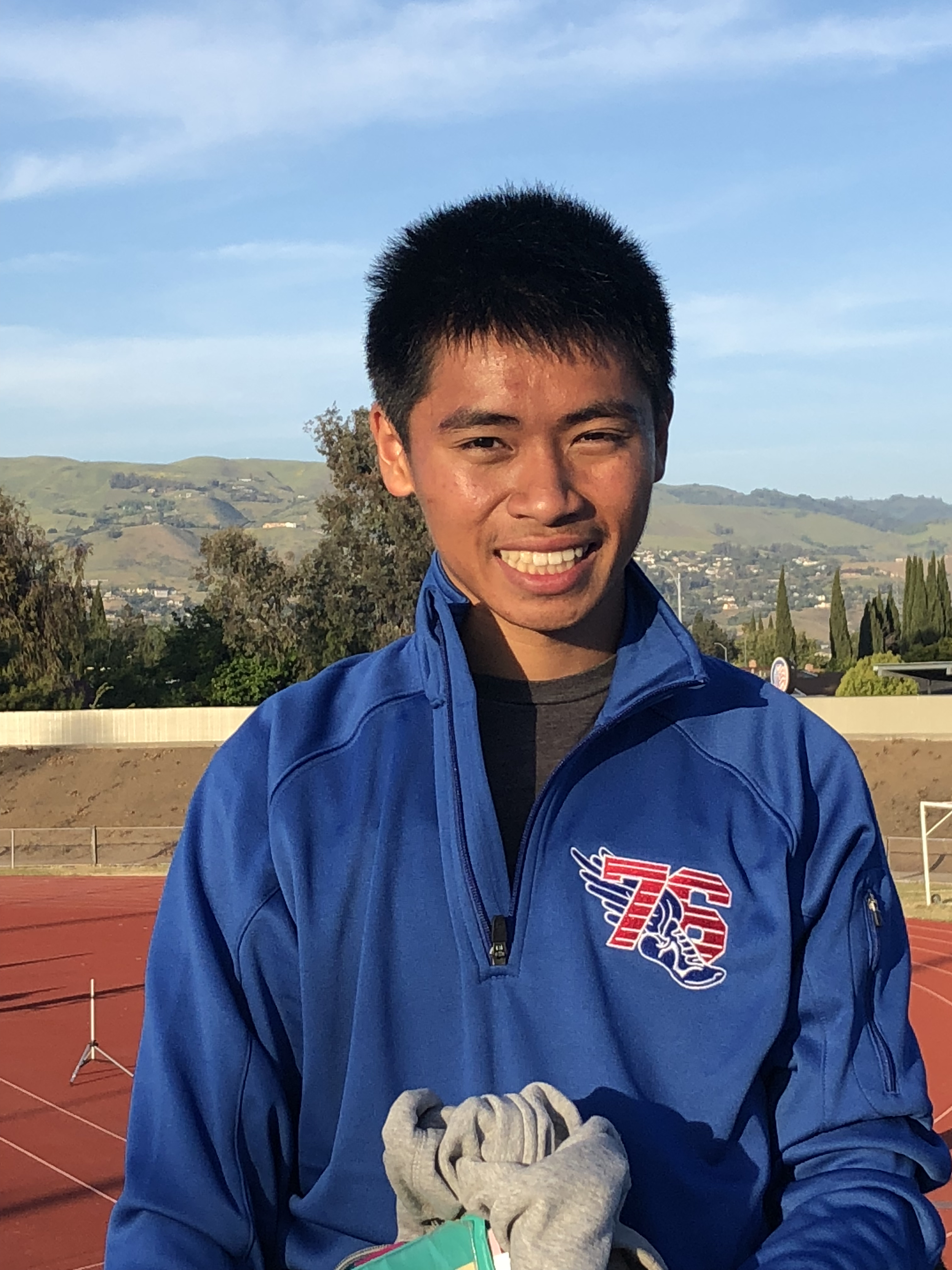 A young man with short black hair is smiling and standing outdoors on a track field, wearing a blue jacket with a logo on the chest, and holding a pair of gray gloves. The background shows hills, trees, and a cityscape under a partly cloudy sky.