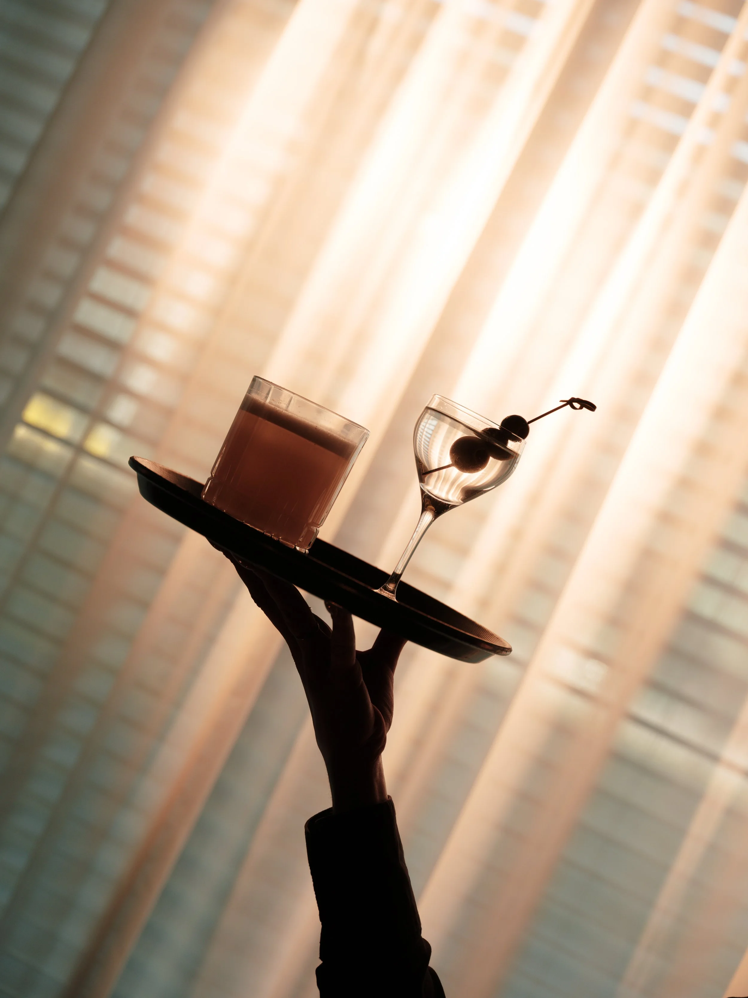 A person holding a black tray with a shot glass filled with a pink drink and a smaller glass of dark cherries on a skewer. Background features window blinds with sunlight filtering through.
