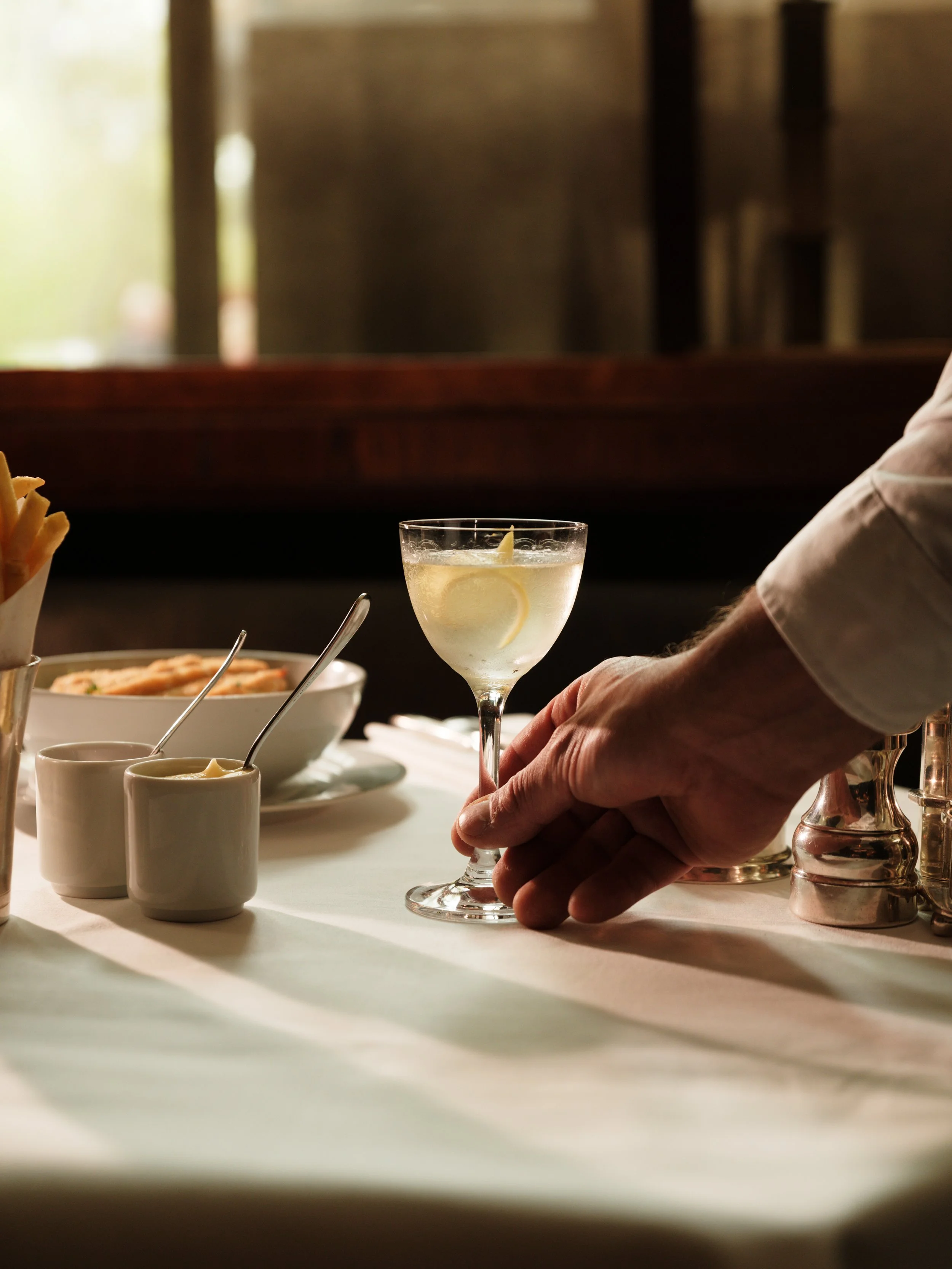 A person holding a cocktail glass with a lemon slice, on a table with bowls of dipping sauces and a basket of breadsticks, in a restaurant setting.