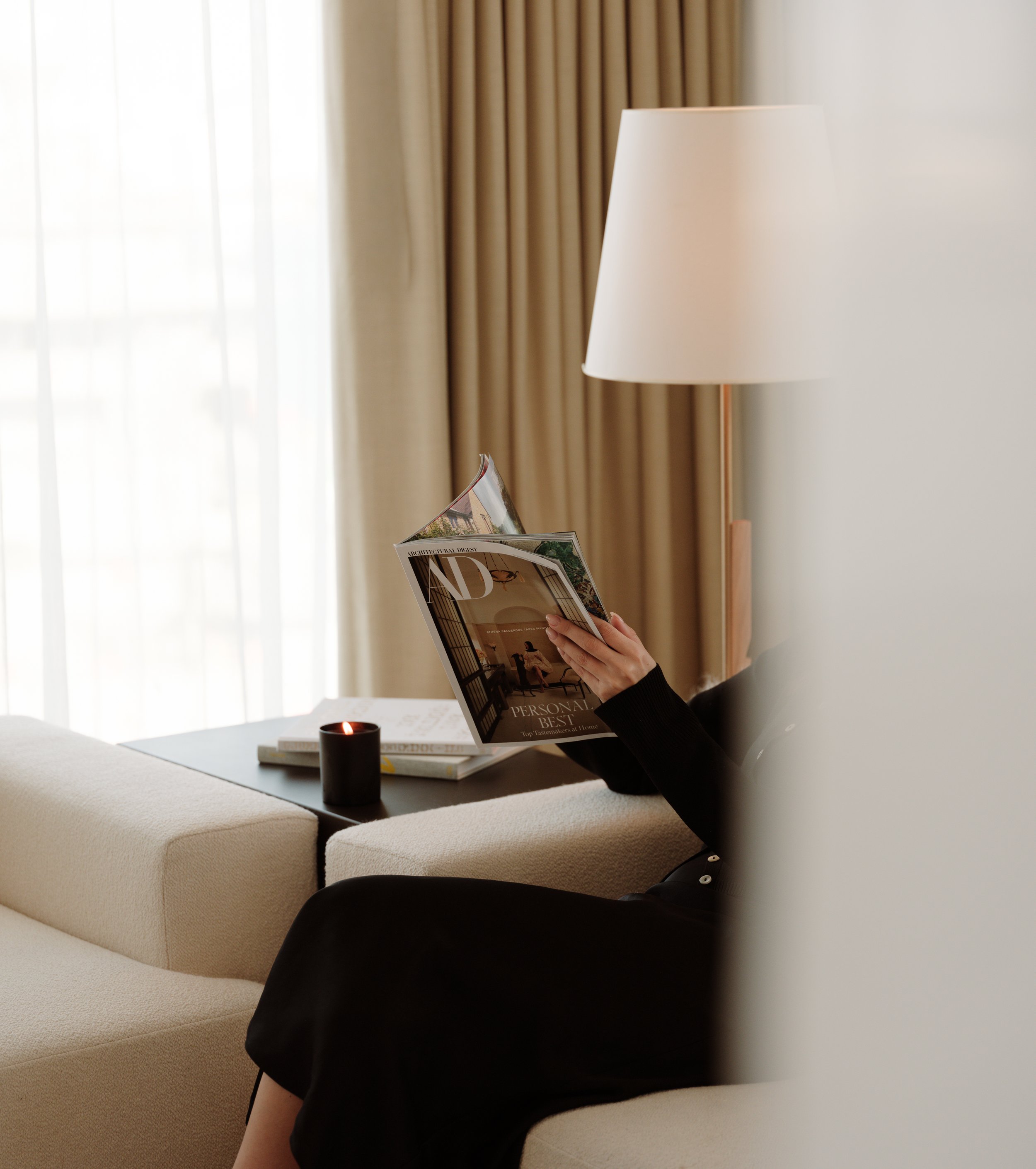 Person sitting on a beige sofa reading a magazine, with a black candle on the table, in a room with beige curtains and a window.