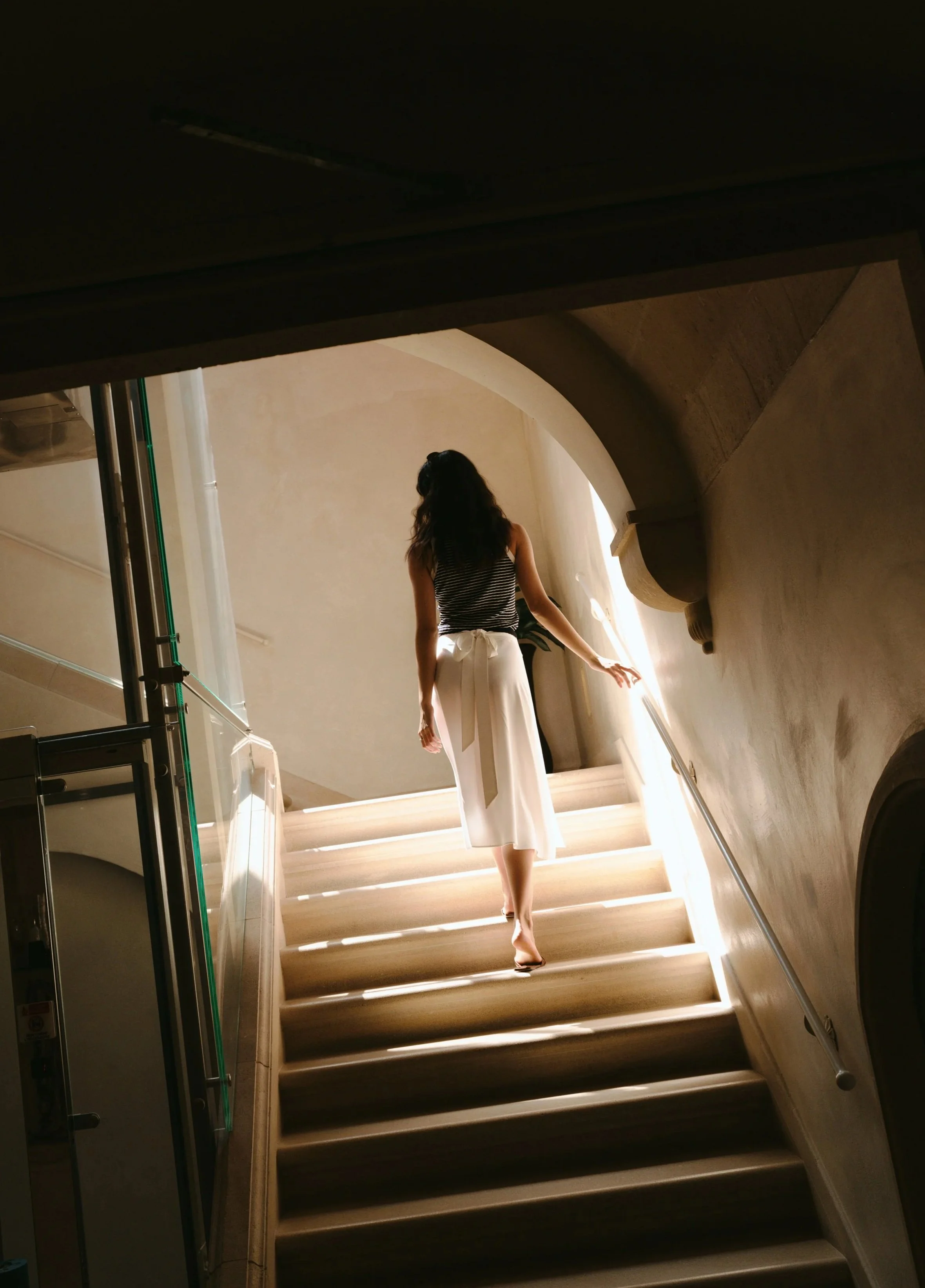 A woman with long hair descending a staircase in a building, illuminated by natural light from a window.