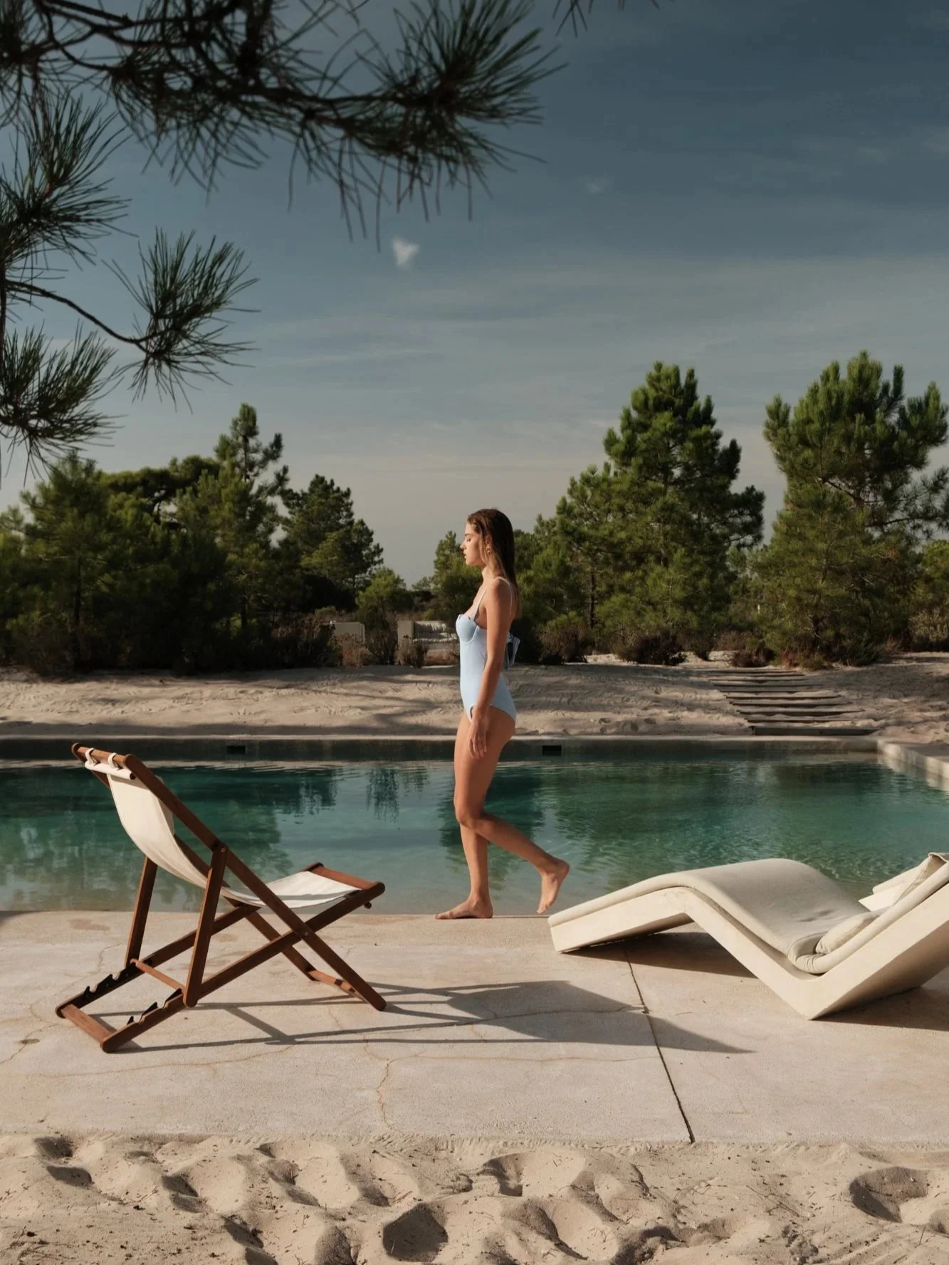 A woman in a blue swimsuit walking along the edge of a swimming pool at a sandy beach. There are two empty lounge chairs, one upright and one reclined, on the concrete deck. In the background, pine trees and a cloudy sky are visible.