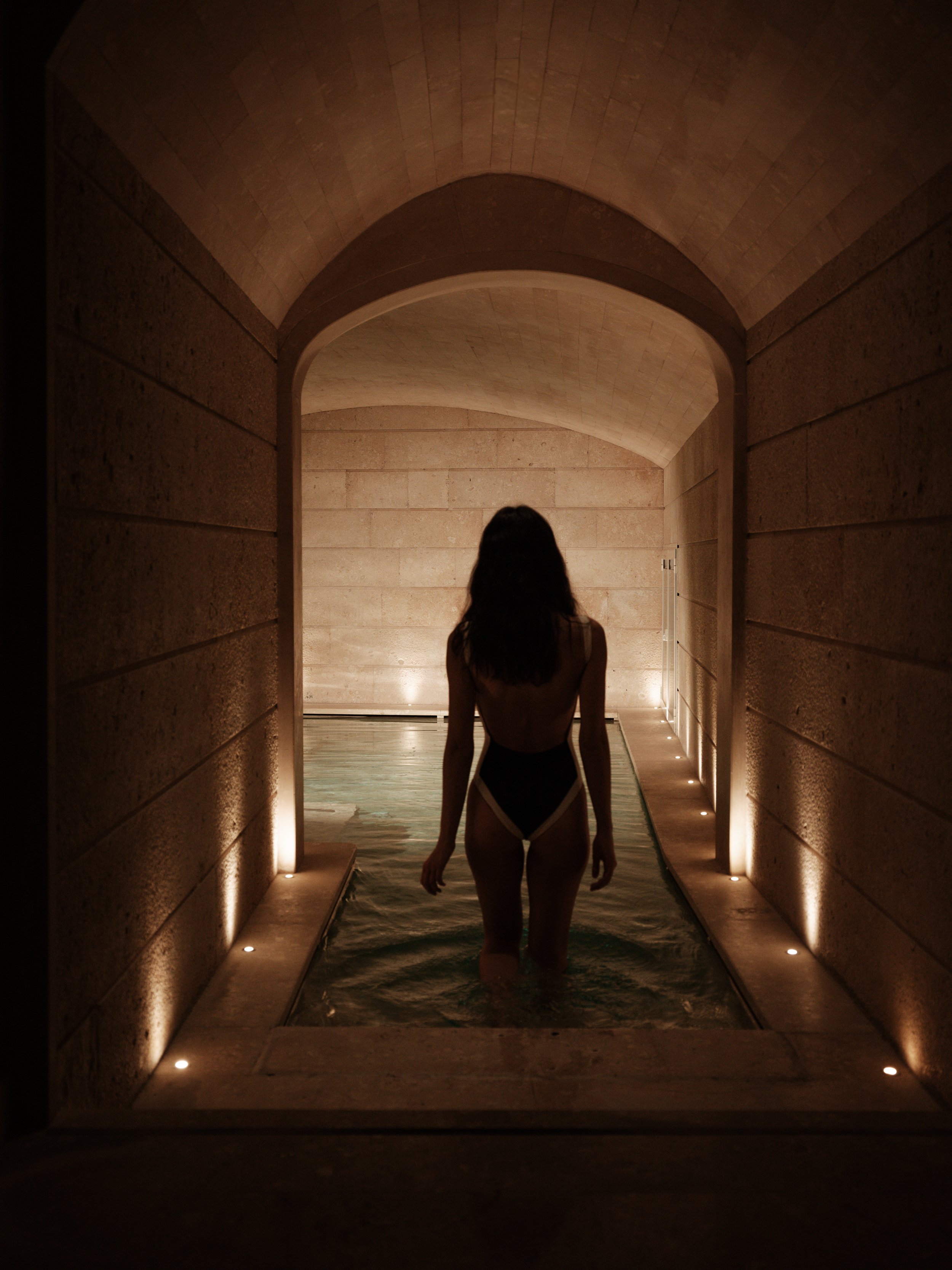 A woman with long hair stands in a dimly lit indoor pool area, facing away from the camera, with arches and wall lights illuminating the space.