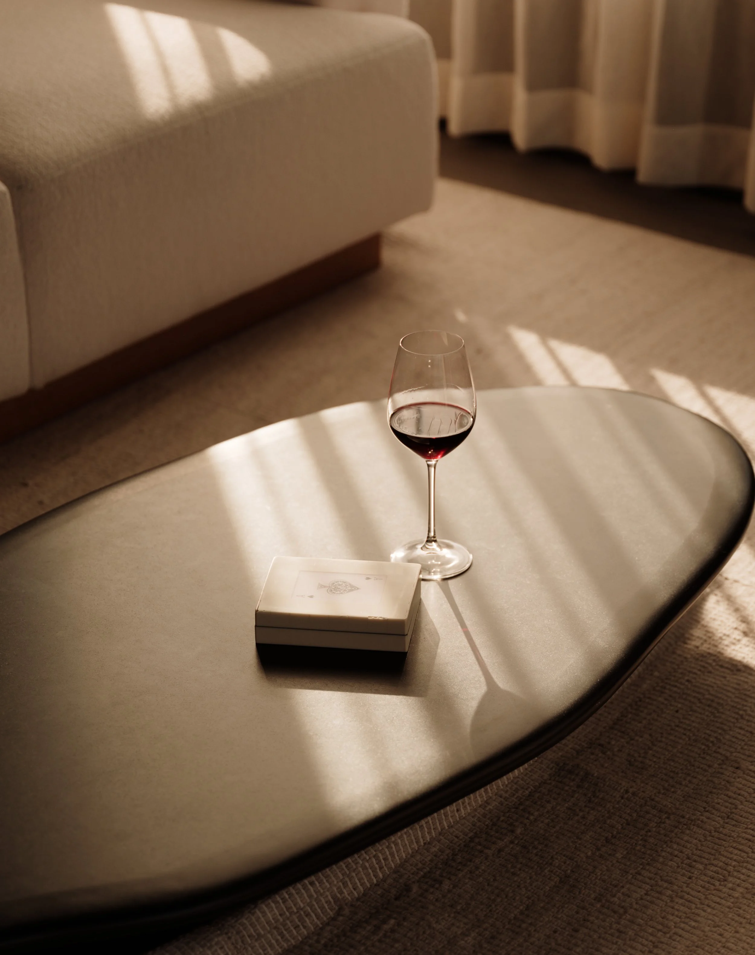 A table with a glass of red wine, a closed deck of playing cards, and a white cloth underneath the cards, in a room with beige carpet and a curved couch and curtains in the background.
