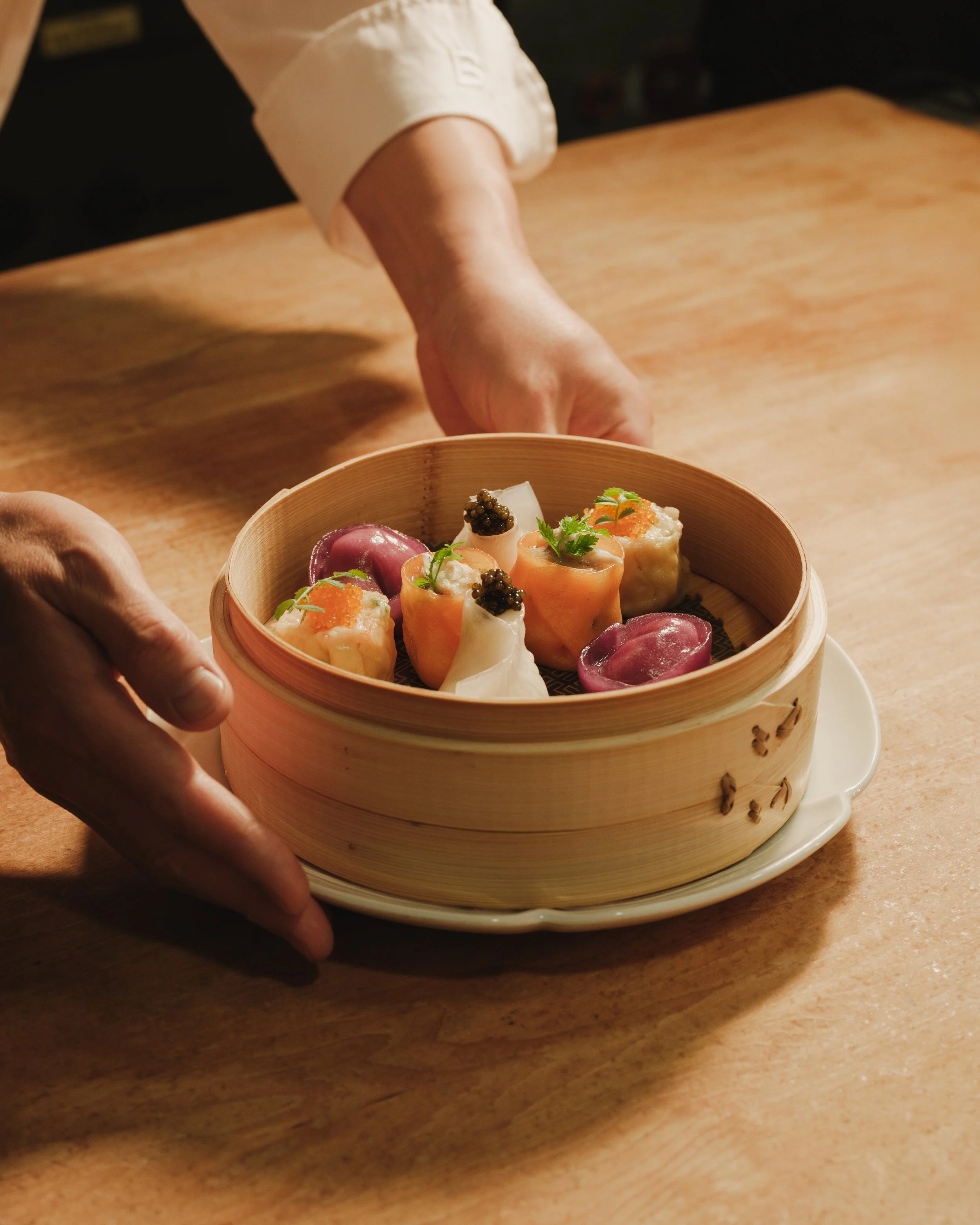 Close-up of a person placing a bamboo steamer filled with assorted sushi on a white plate on a wooden table.
