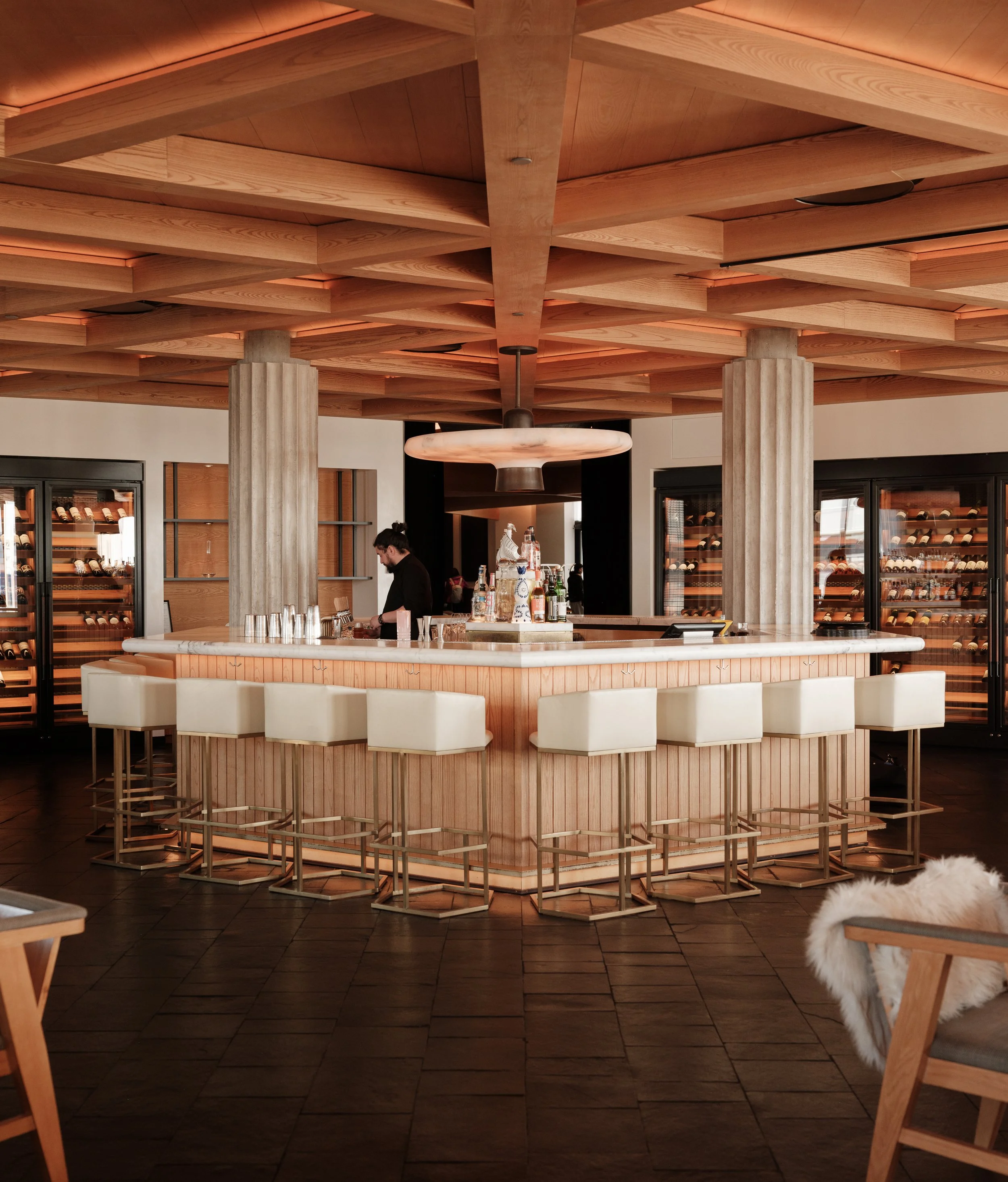 Interior of a modern restaurant or bar with a semi-circular bar counter made of light wood and a marble top. Behind the bar, shelves with wine bottles are visible. Two tall, fluted columns support a wooden ceiling with geometric patterns. A bartender