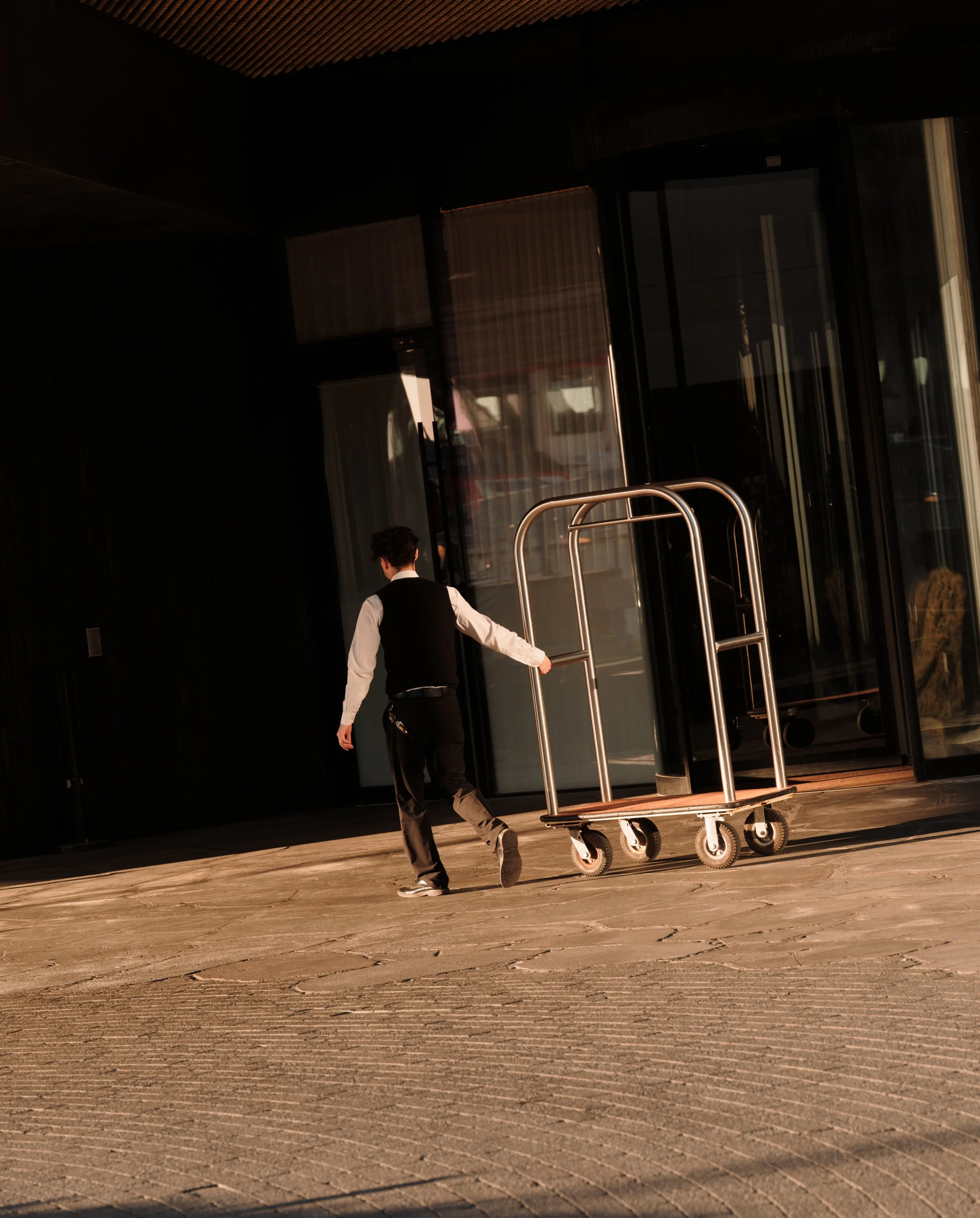 A man in formal attire, wearing a black vest and white shirt, is pushing a luggage cart outside a modern building with glass doors and large windows. The scene is illuminated by natural light, casting shadows on the textured pavement.