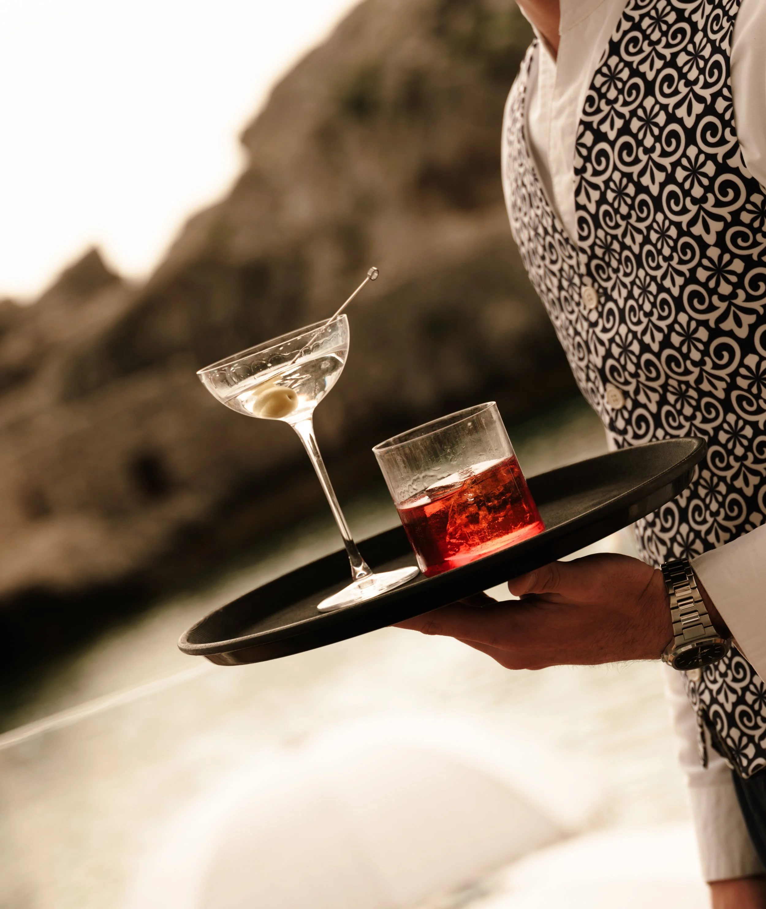 Person dressed in a patterned vest and white shirt holding a tray with a martini glass containing an olive and clear liquid, and a glass of red liquid, against a blurred outdoor background.