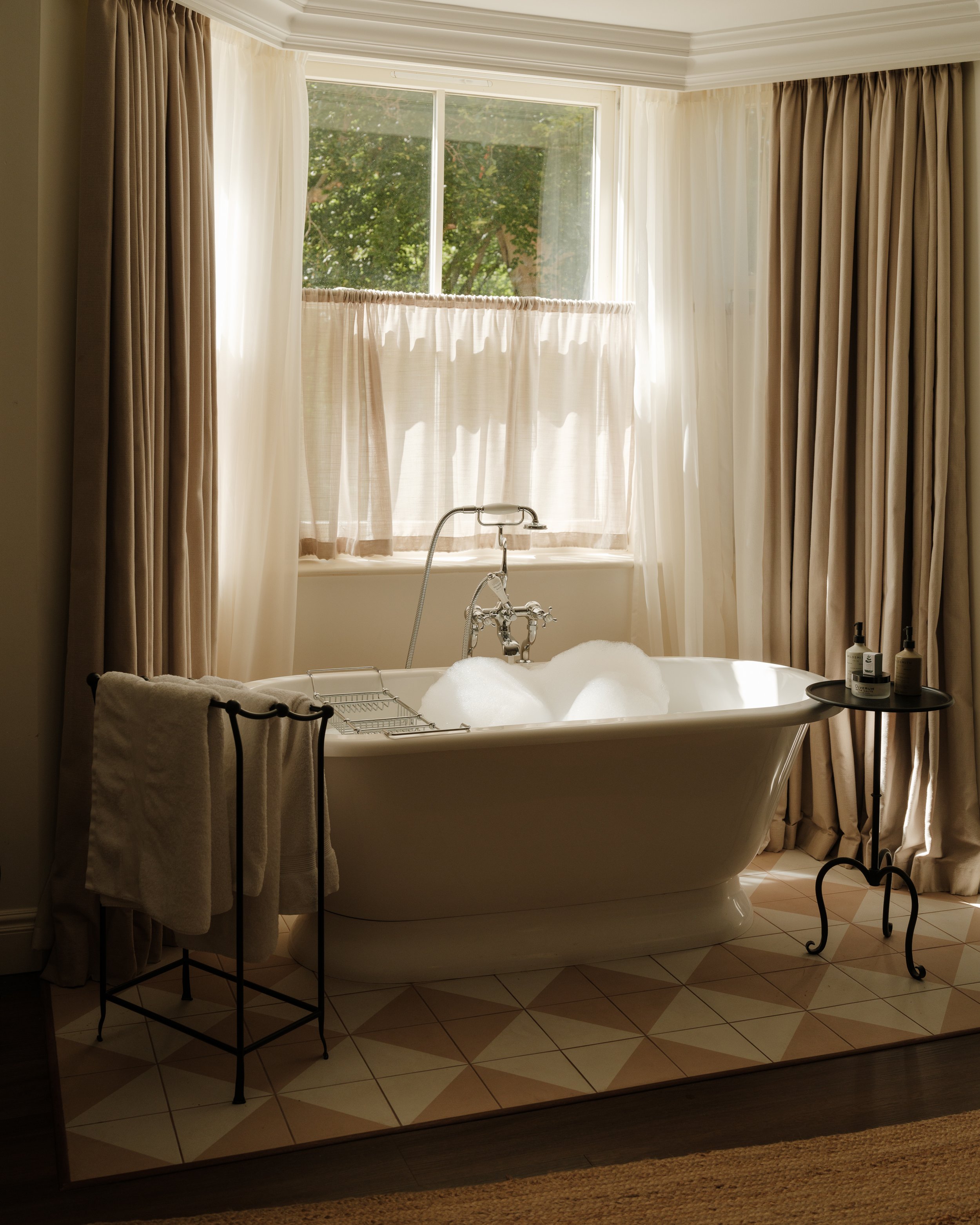 A bathroom with a white clawfoot bathtub filled with bubbles, located in front of a large window with sheer and blackout curtains, and a small black table with lotion bottles on it. There is a towel rack with a folded towel in front of the bathtub an