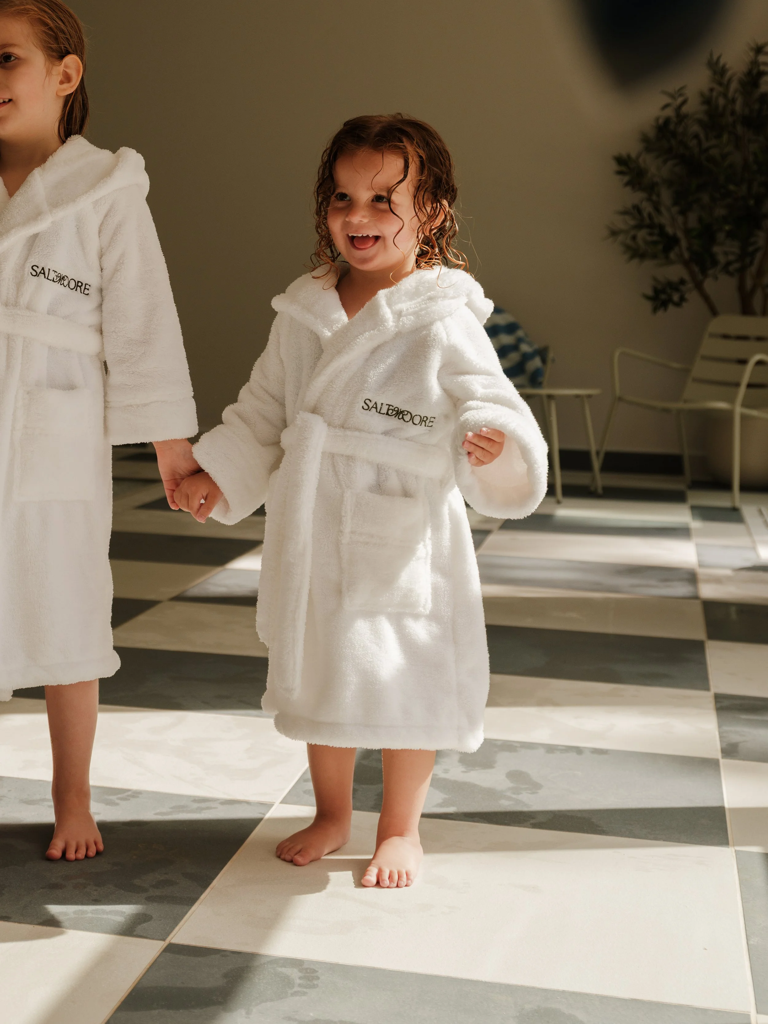 Two young children in white robes standing on a checkered floor, with the child on the right smiling and the feet of another child visible on the left.