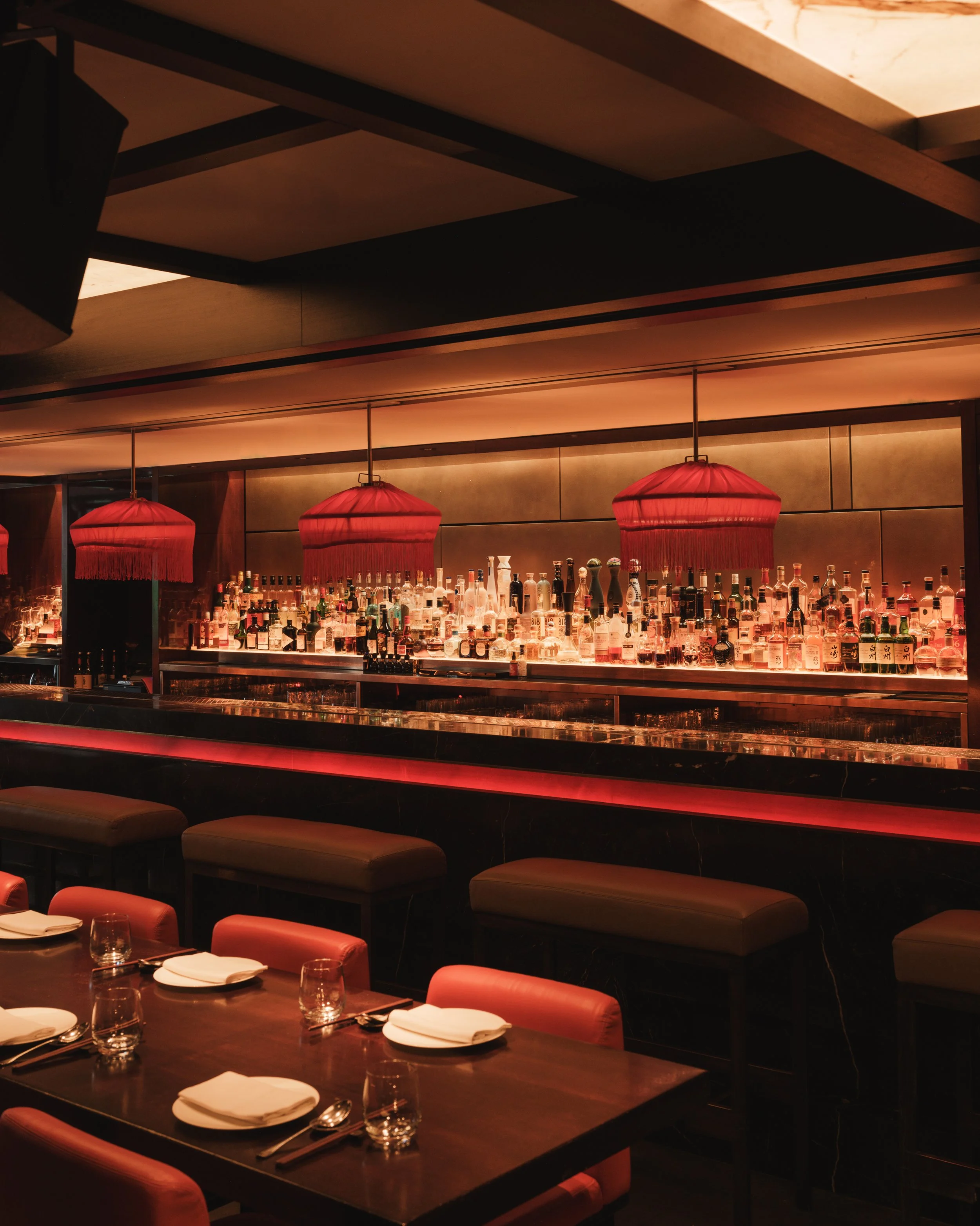 Interior of a restaurant or bar with a backlit bar counter stocked with various bottles of alcohol, two red hanging pendant lights, and a dark wood dining table set with napkins, glasses, and silverware.