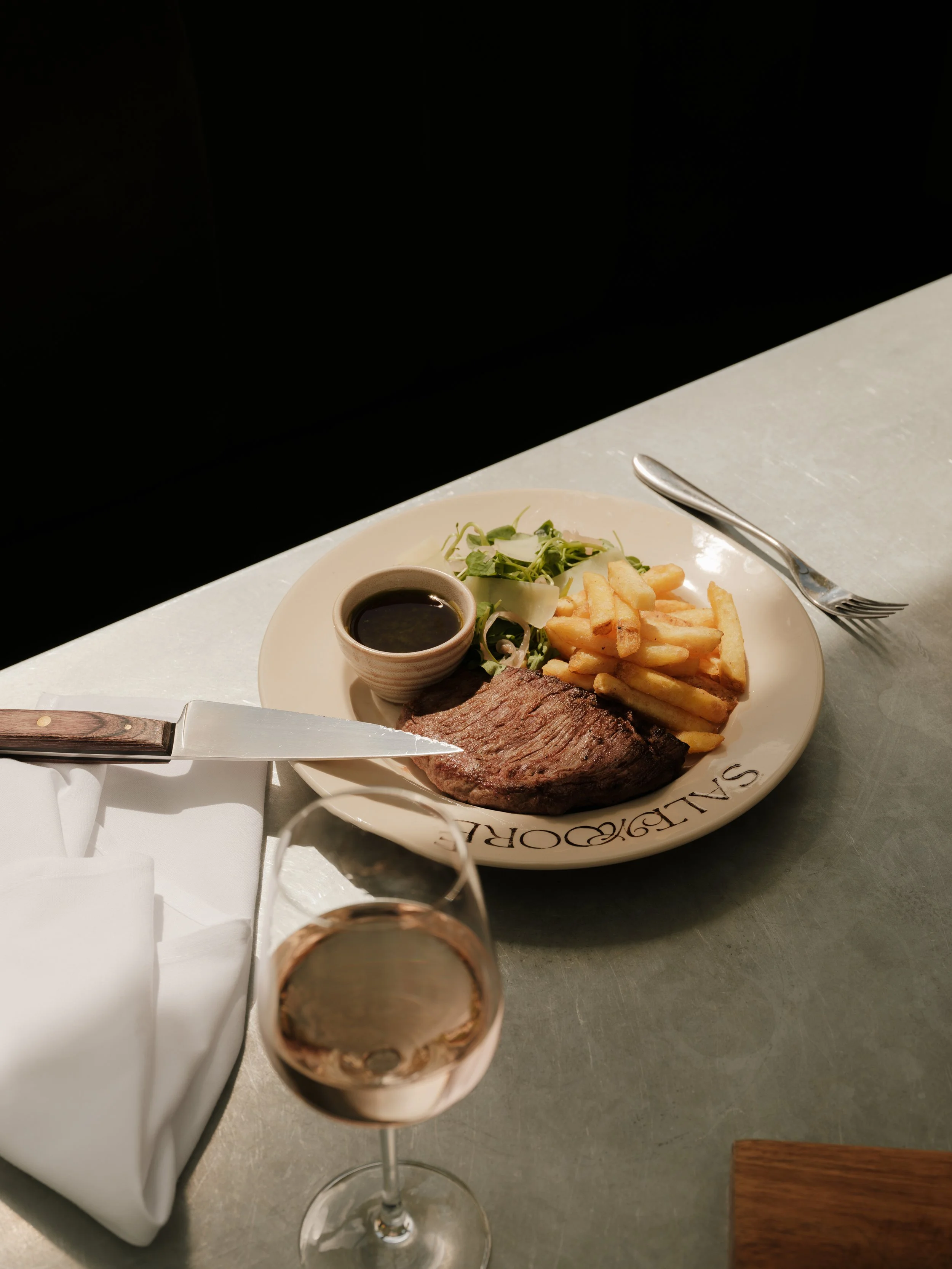 A meal consisting of a grilled steak, French fries, mixed green salad, and a small bowl of dipping sauce on a white plate. Also visible is a glass of red wine and a knife on a white napkin on a table.
