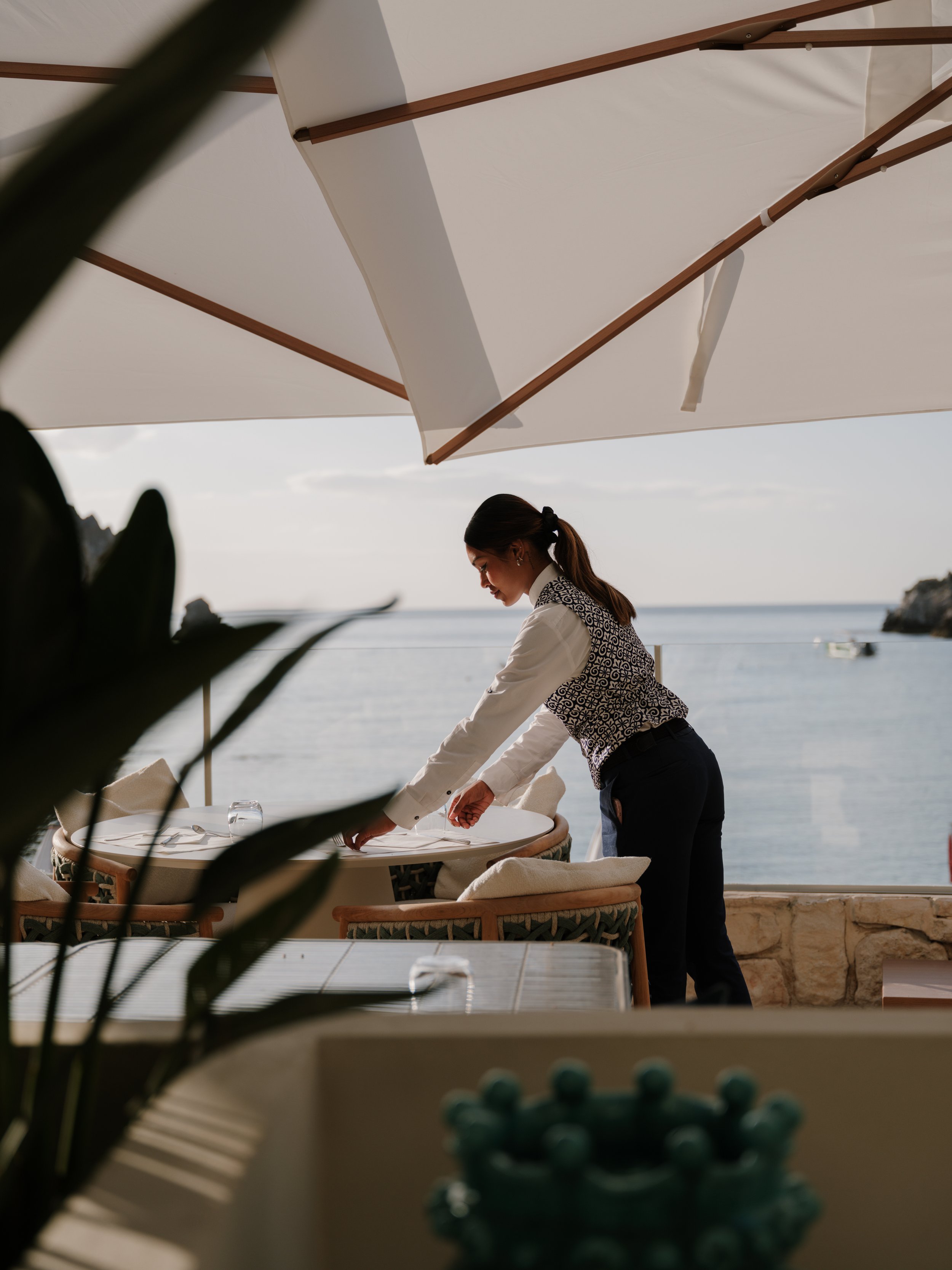 A woman with dark hair tied back, wearing a patterned vest and white shirt, setting a table at an outdoor restaurant near the water under a large sun umbrella.
