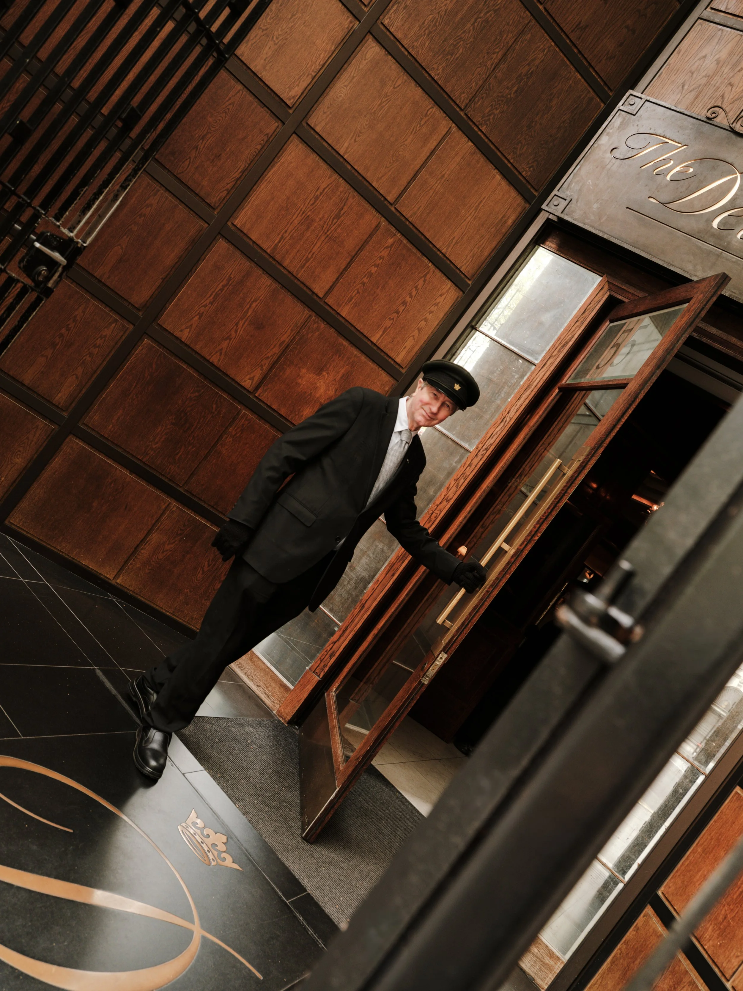 A hotel bellhop in a black uniform and cap standing in an elegant hotel lobby next to a wooden display wall, visible through a glass door.