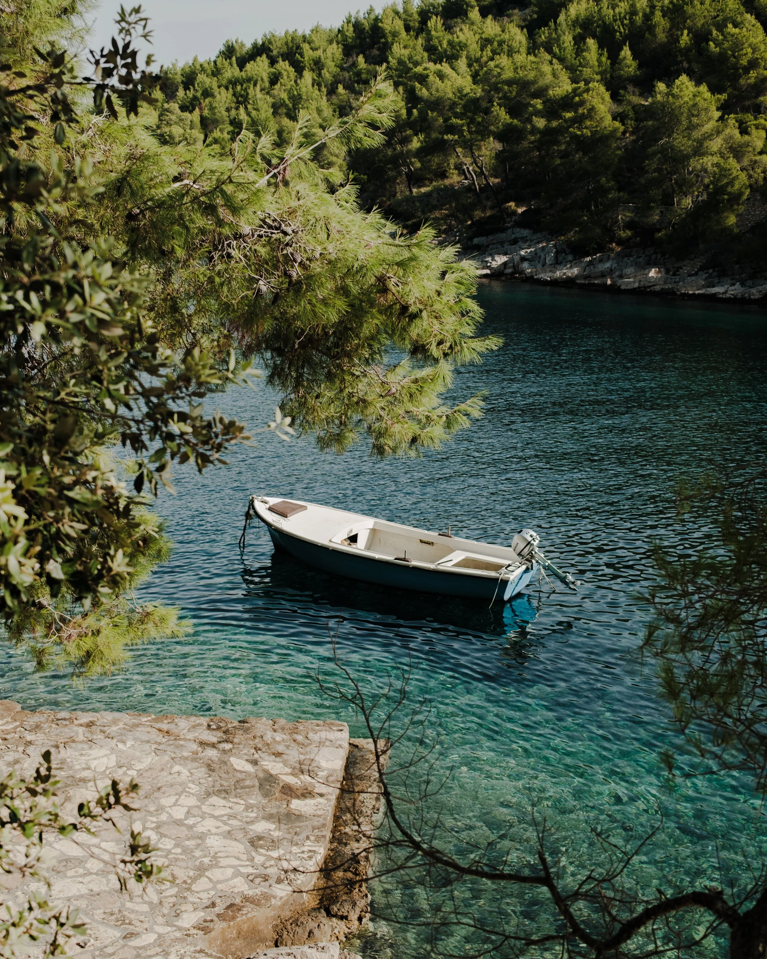 A small white boat with an outboard motor floating on clear blue-green water, surrounded by rocky shoreline and dense green trees.