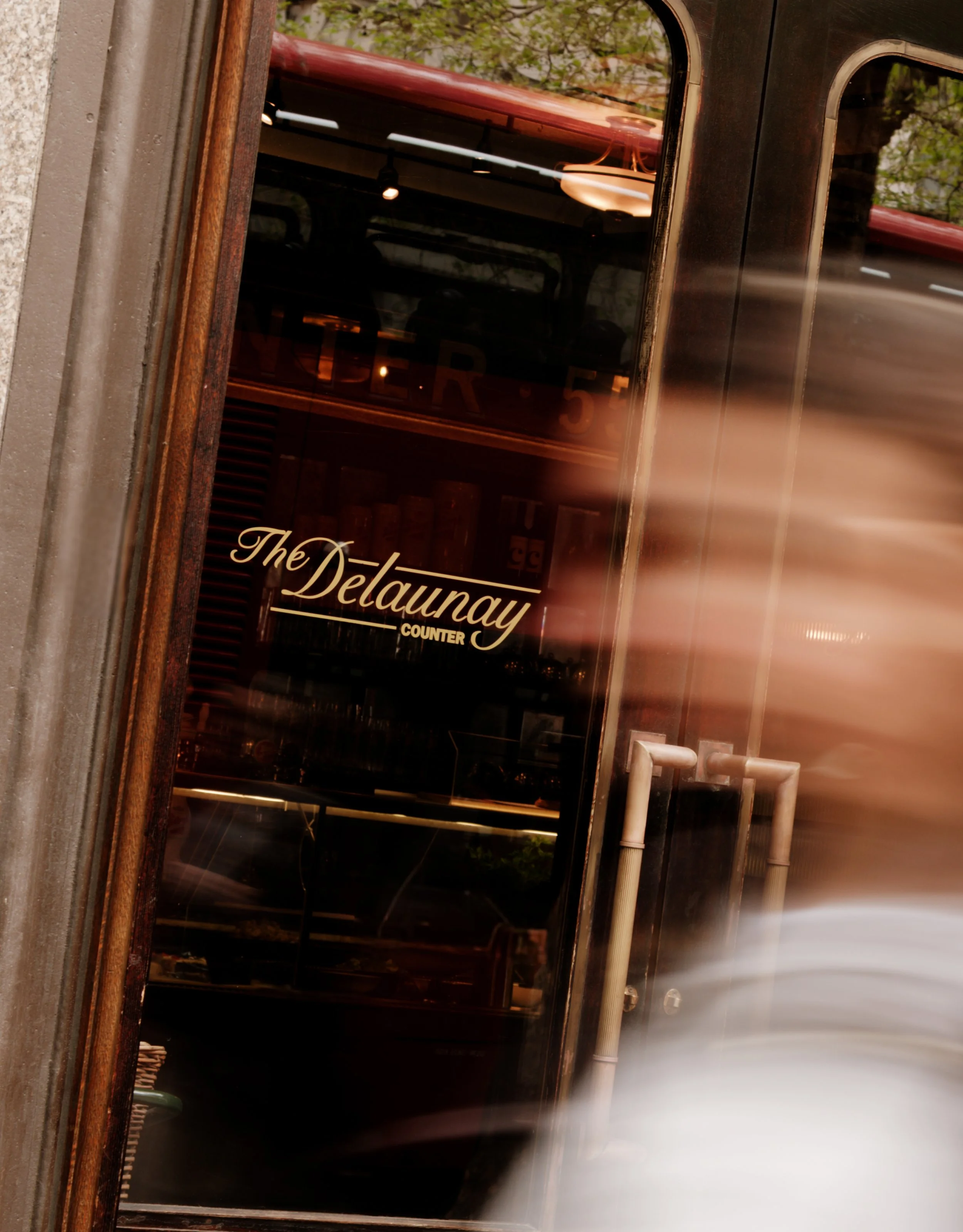 A storefront window with the sign 'The Delaunay' and part of the word 'Counter'. Reflection of a tree and street scene in the glass. Piping and sidewalk in the foreground.