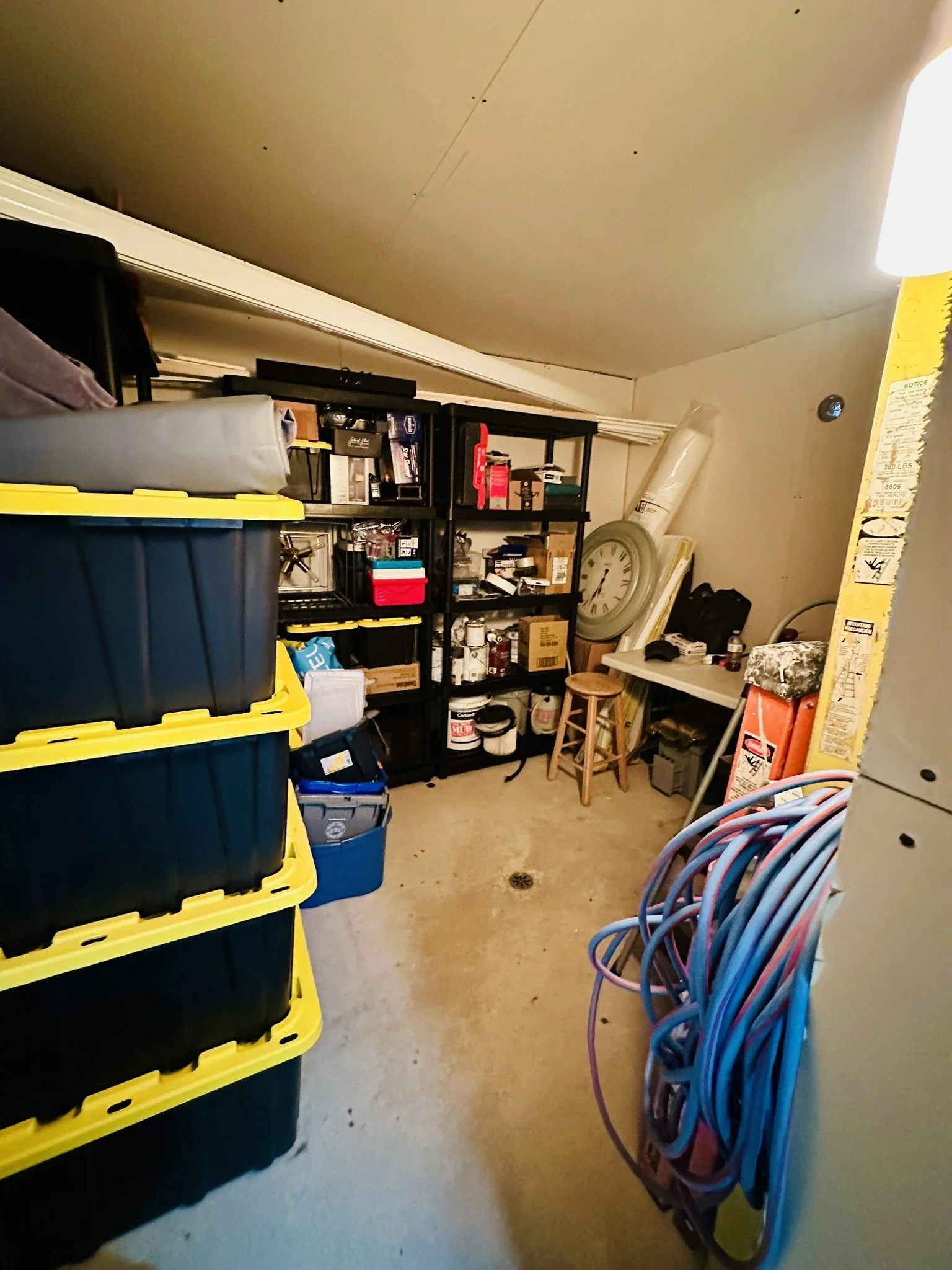 Laundry or storage room with black shelves filled with boxes, bottles, and miscellaneous items; blue and purple cables hanging on the right; plastic storage bins on the left; white ceiling and beige floor with a drain.