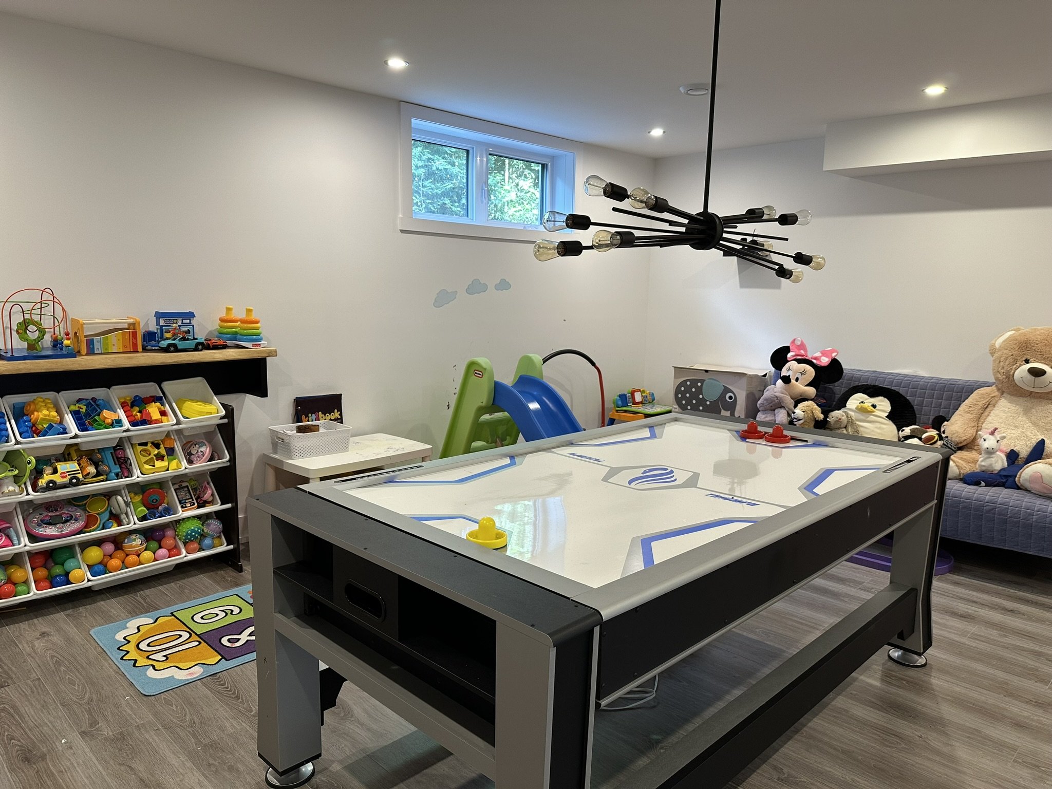 A basement game room with an air hockey table, plush toys including Minnie Mouse and a teddy bear, toy storage with bins filled with colorful toys, a small slide, and a window with a view of greenery outside. There are toys on a shelf and a cartoon-themed rug on the floor.