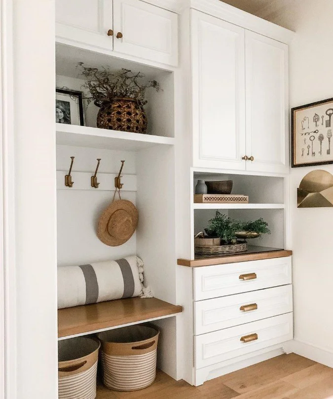 Built-in white storage with open shelves, drawers, and hooks, decorated with baskets, a hat, a rolled towel, and decorative items, with a framed artwork of keys on the wall.