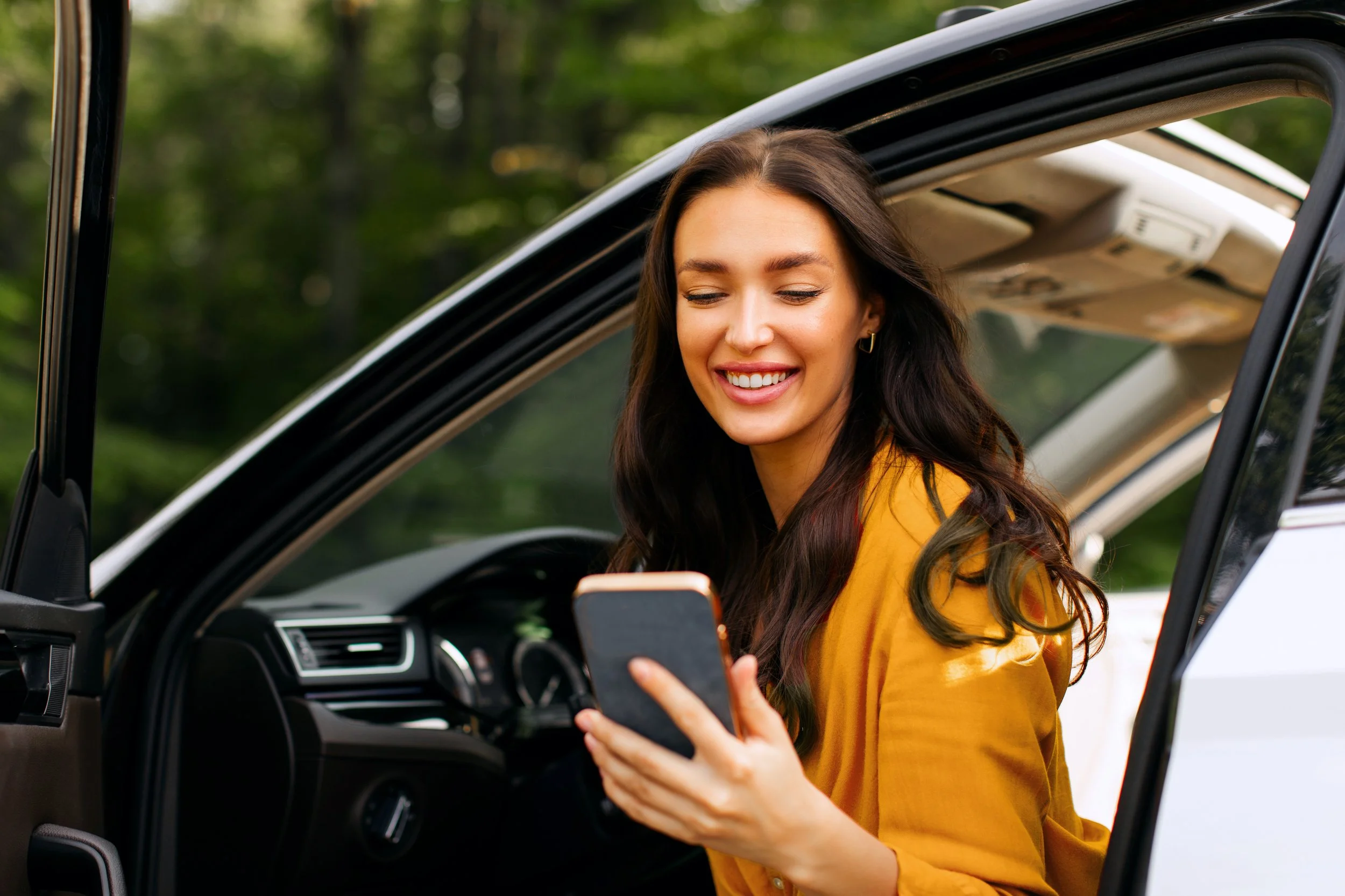 A young woman with long dark hair smiling and looking at her phone while sitting in the driver's seat of a car.