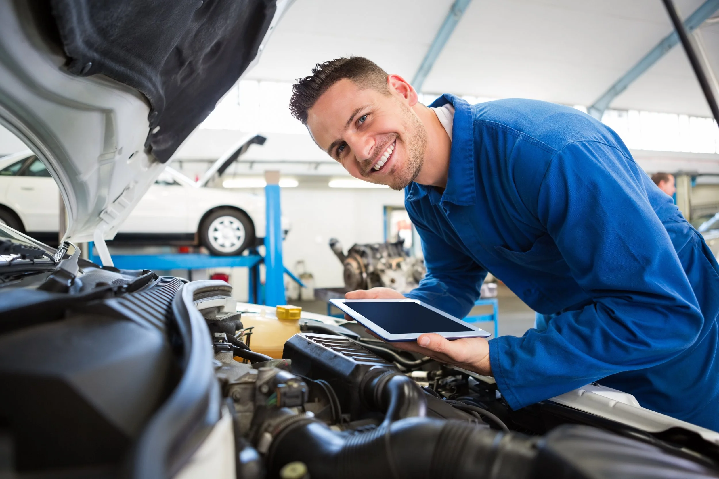 A mechanic in a blue uniform working under the hood of a car in an auto repair shop, holding a tablet and smiling at the camera.
