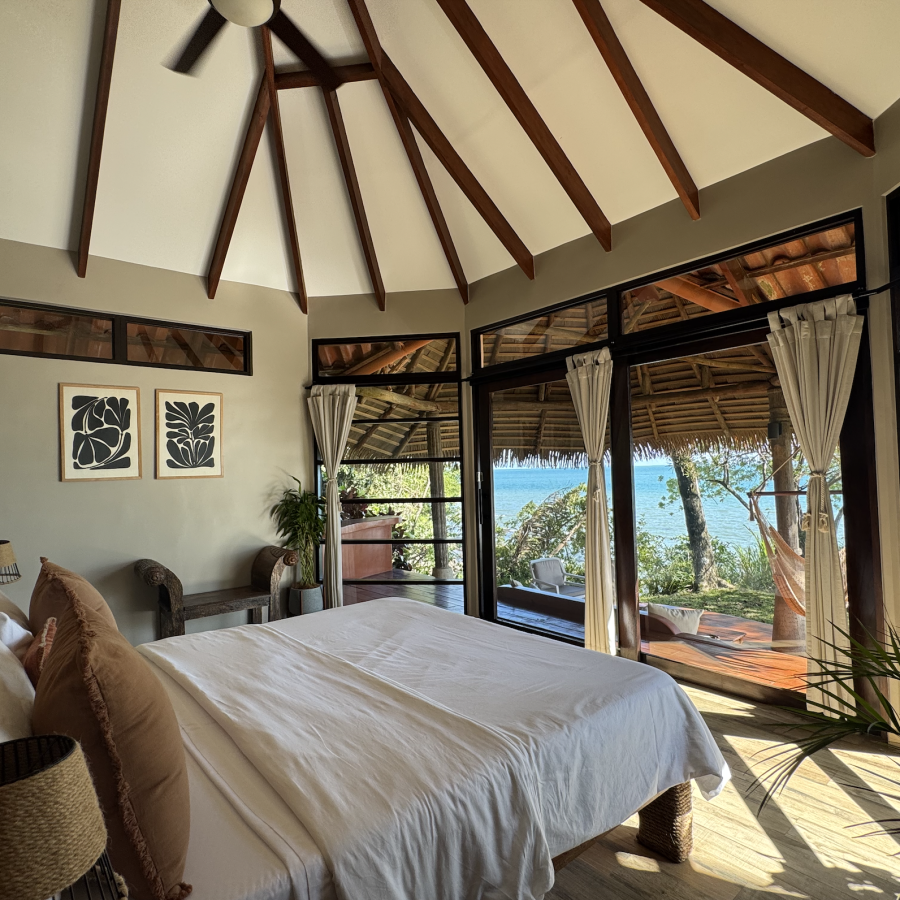 Bright Private bedroom with a vaulted ceiling, wooden beams, and large sliding glass doors opening to a balcony with a view of trees and water.