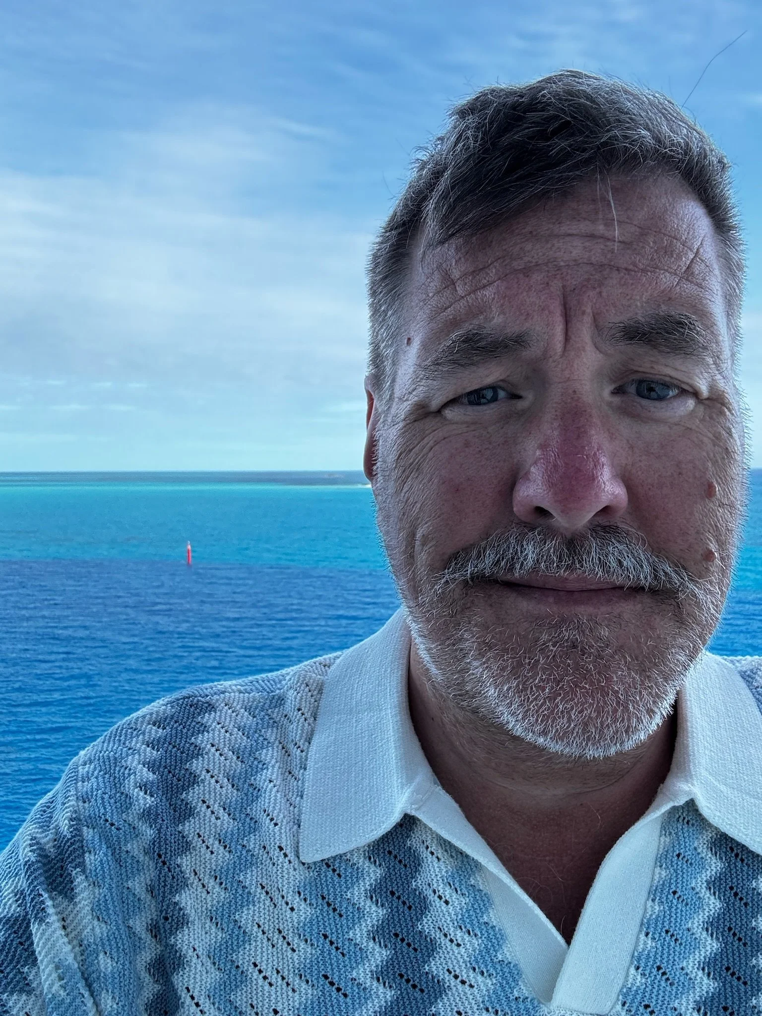 A man with gray hair, mustache, and beard taking a selfie near a body of water, with a blue sky and horizon in the background.