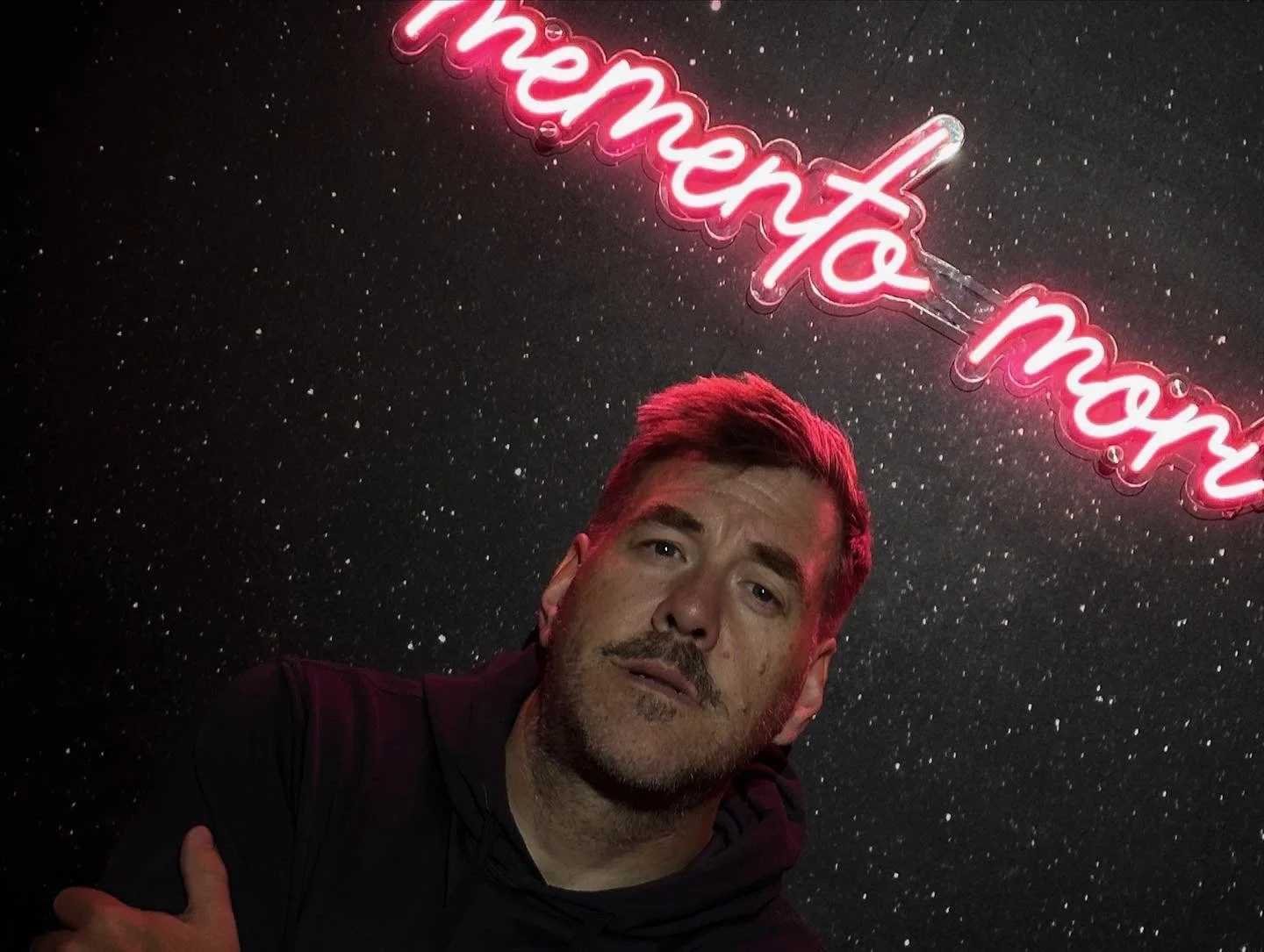 Man with short brown hair and a beard standing in front of a black starry background with a pink neon sign that reads 'moment to moment' hanging above him.
