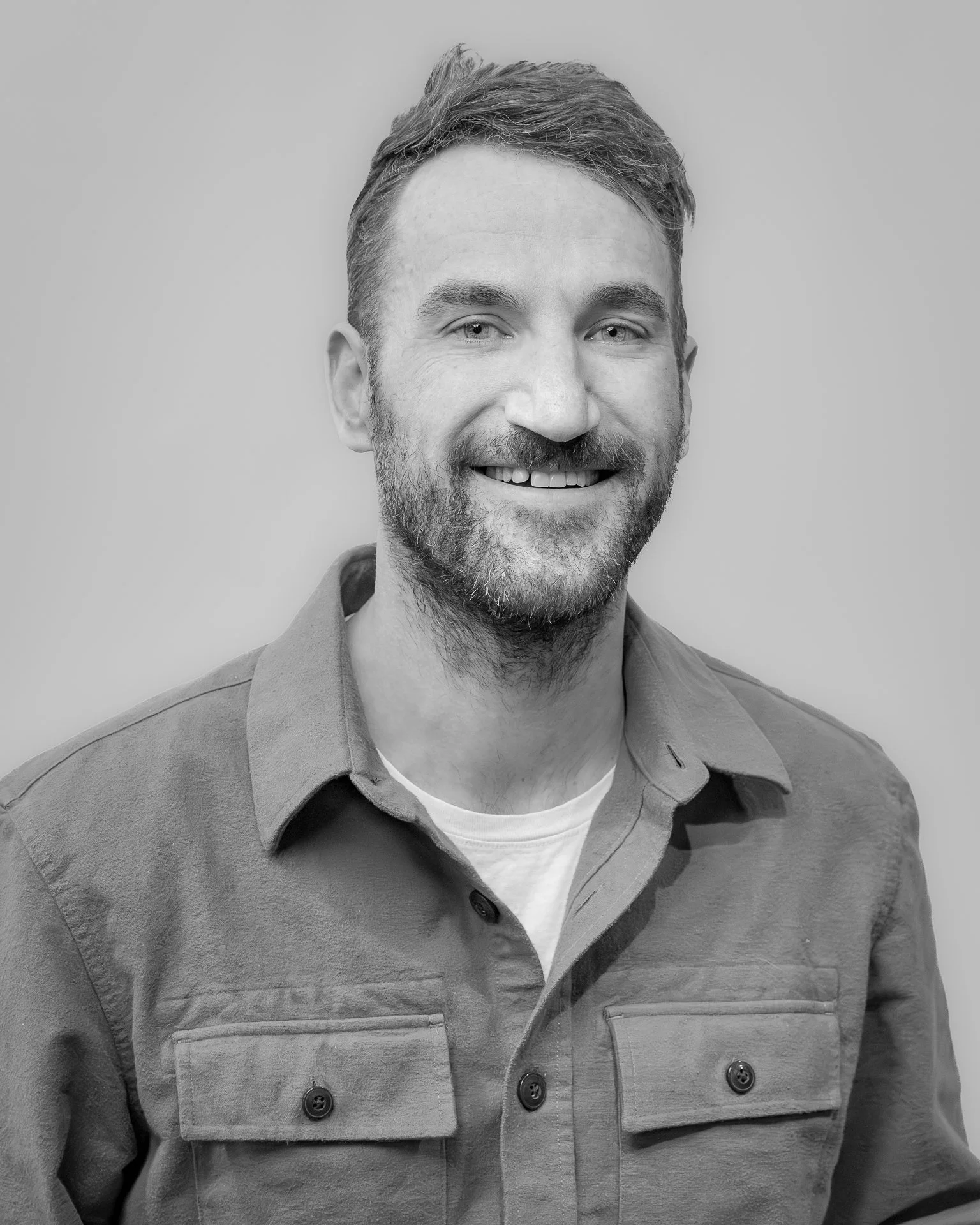 Black and white portrait of a smiling man with short hair and a beard, wearing a button-up shirt over a t-shirt.