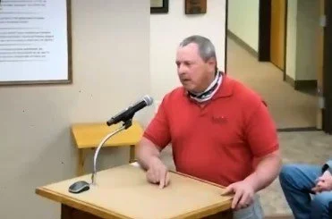 Man testifying at a podium with a microphone in a room with beige walls.