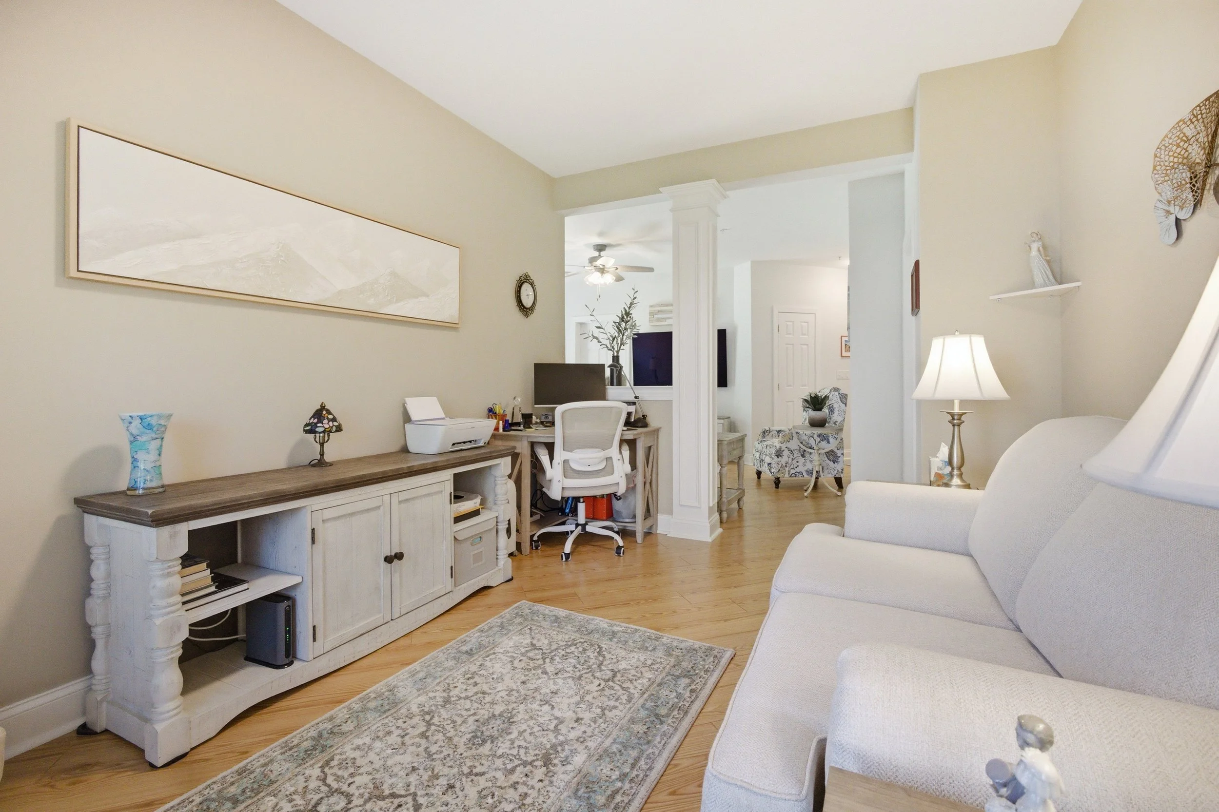A cozy living room with a white armchair, a wooden console table holding a lamp and decorative items, and a desk with a chair and computer. There is a ceiling fan, wall art, and a patterned rug on a wooden floor.
