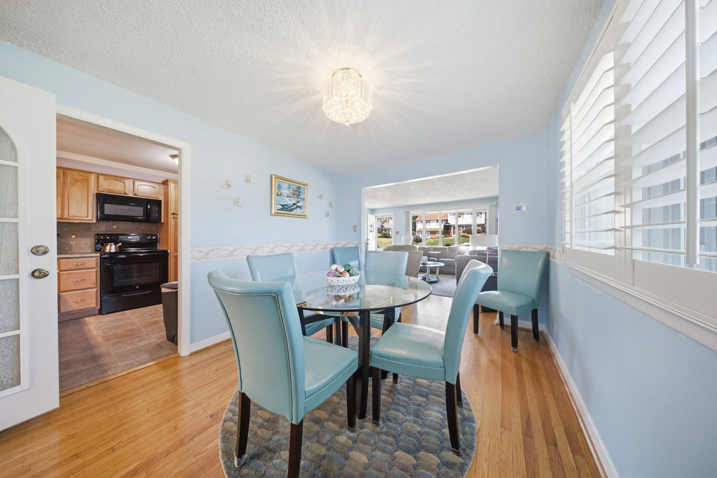 A bright living and dining area with light blue walls, hardwood floors, a glass dining table with blue chairs, a white sofa, and large windows with white plantation shutters, illuminated by a ceiling light fixture.