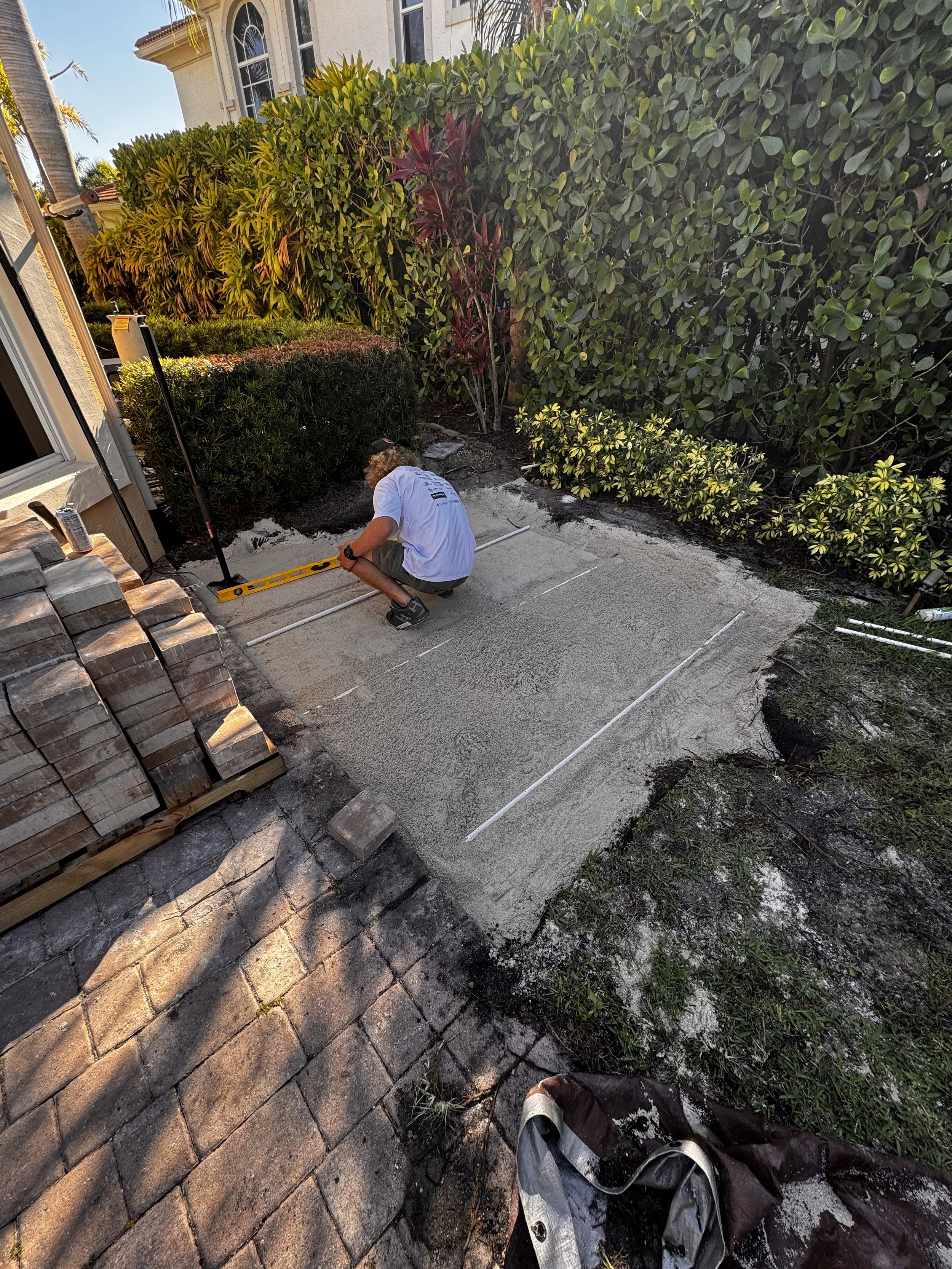 A person working on a concrete patio, using a level and tools, surrounded by bushes and a house.