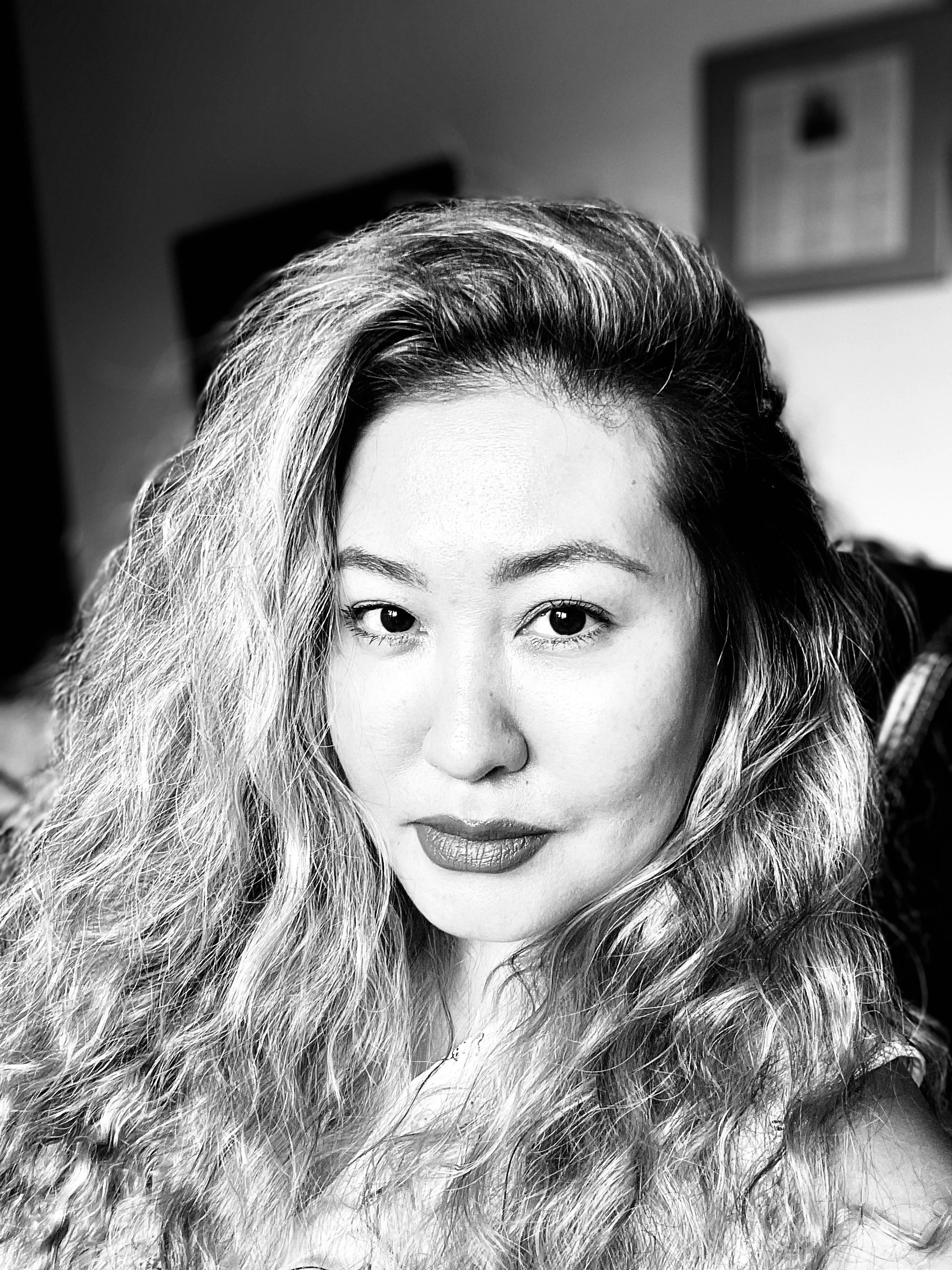 Black and white close-up portrait of a woman with long, wavy hair, looking directly at the camera with a slight smile.